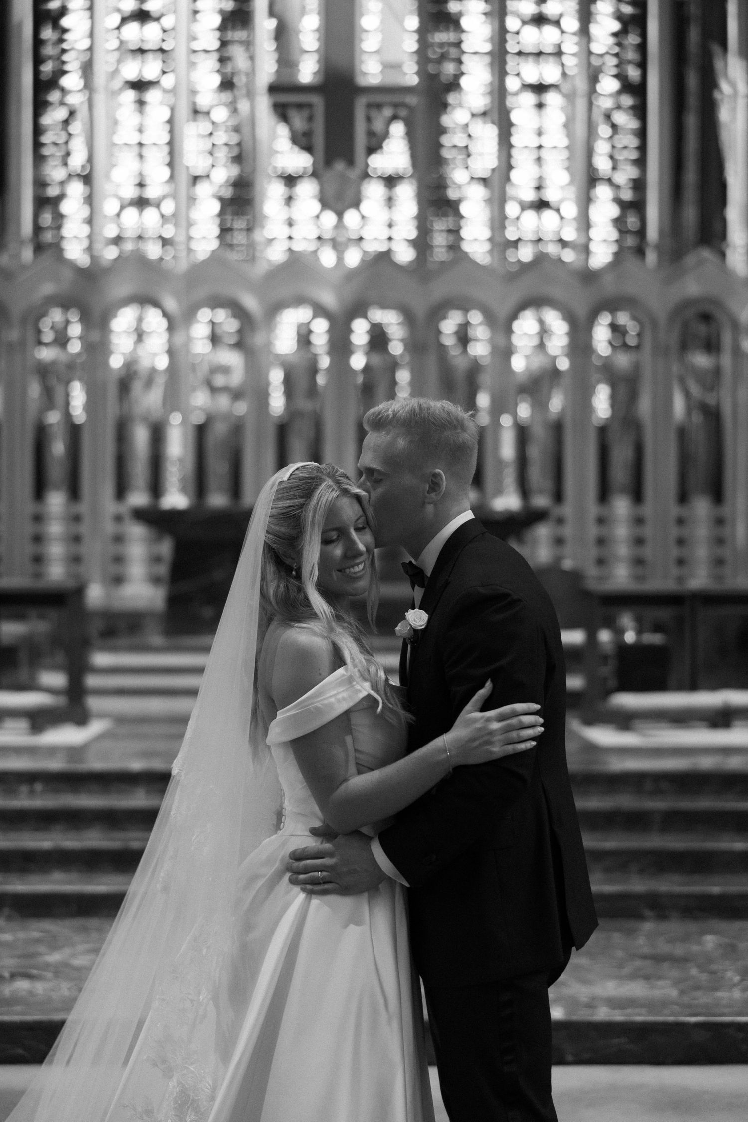 Black and white photo of bride and groom embracing at church altar with stained glass.