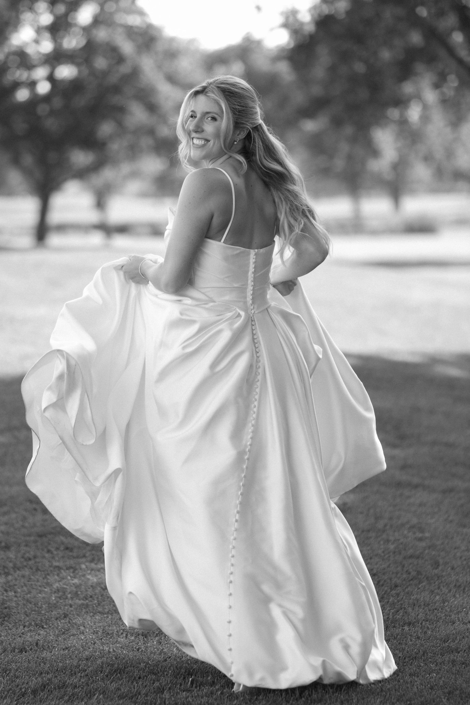 Smiling bride twirling in a flowing white satin wedding gown outdoors in a park, black and white photo.