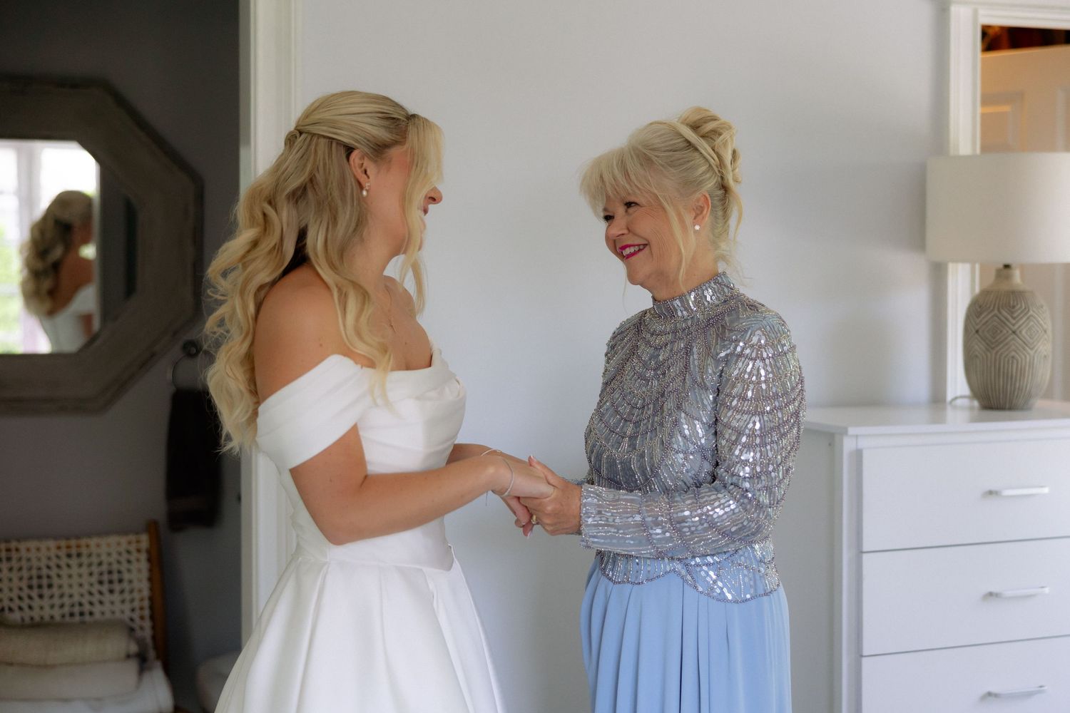 Bride in white off-shoulder gown holding hands with older woman in silver top and blue skirt in bright room.