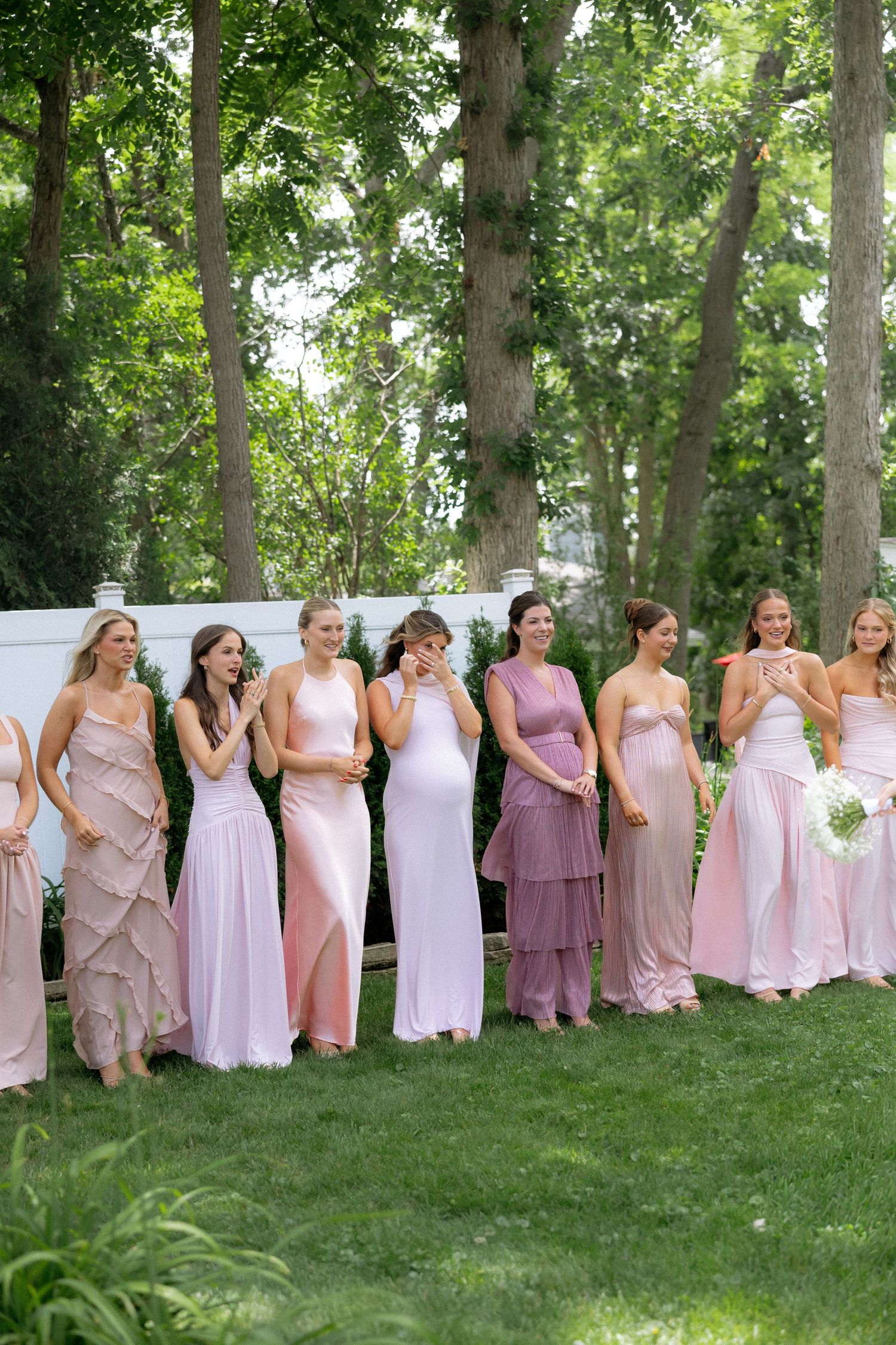 Eight bridesmaids in pink and mauve dresses standing outdoors laughing together.