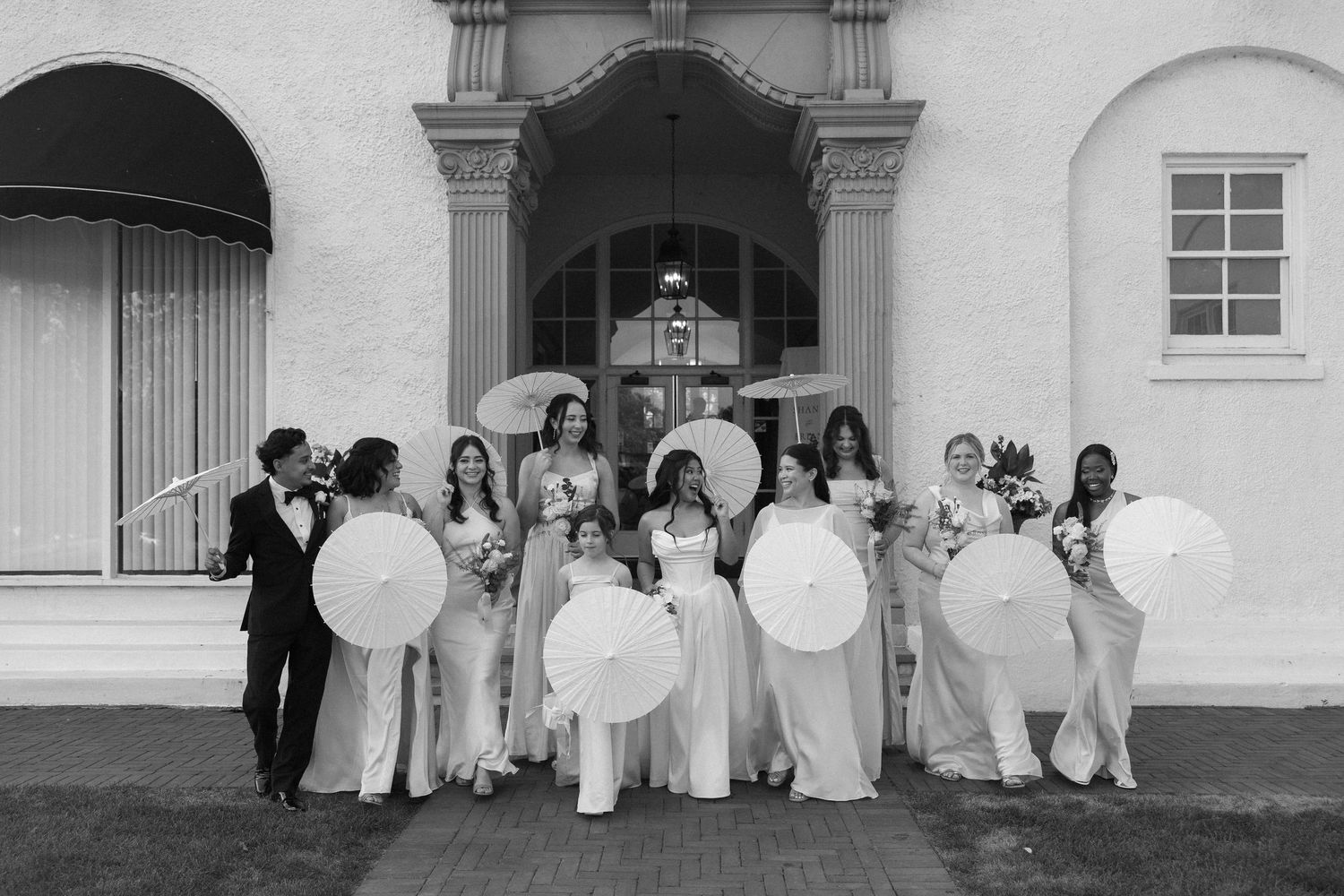 Wedding party poses in black and white photo holding parasols outside elegant white venue.