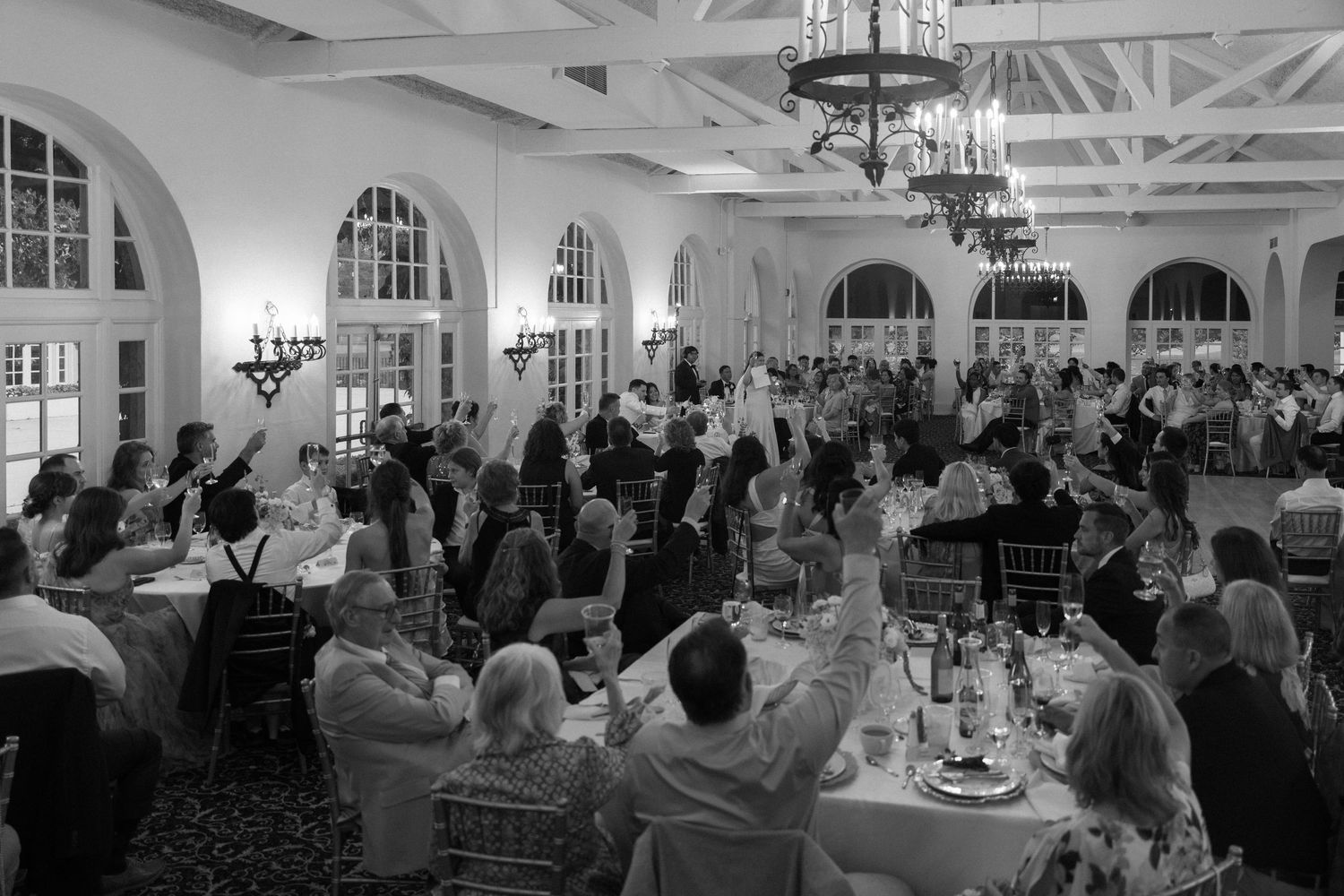 Black and white wide shot of wedding reception guests celebrating in grand ballroom with chandeliers.