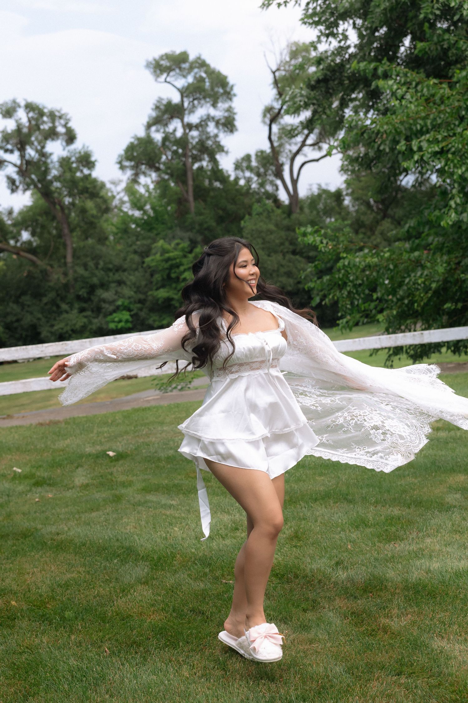Joyful bride twirling in white mini dress and lace cardigan on green lawn with trees in background.