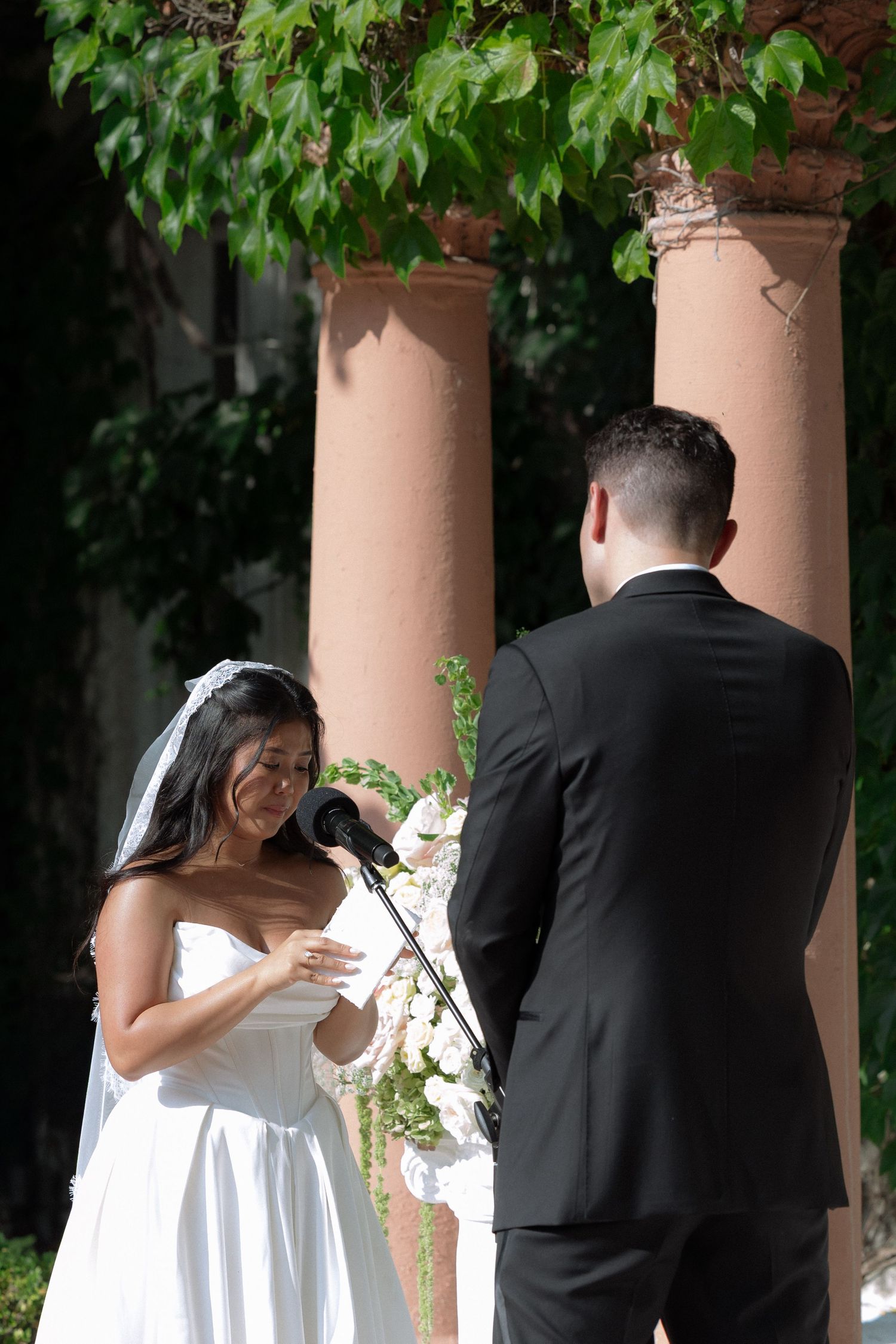 Bride in white dress and veil reads wedding vows at outdoor ceremony with groom in black suit near stone columns.