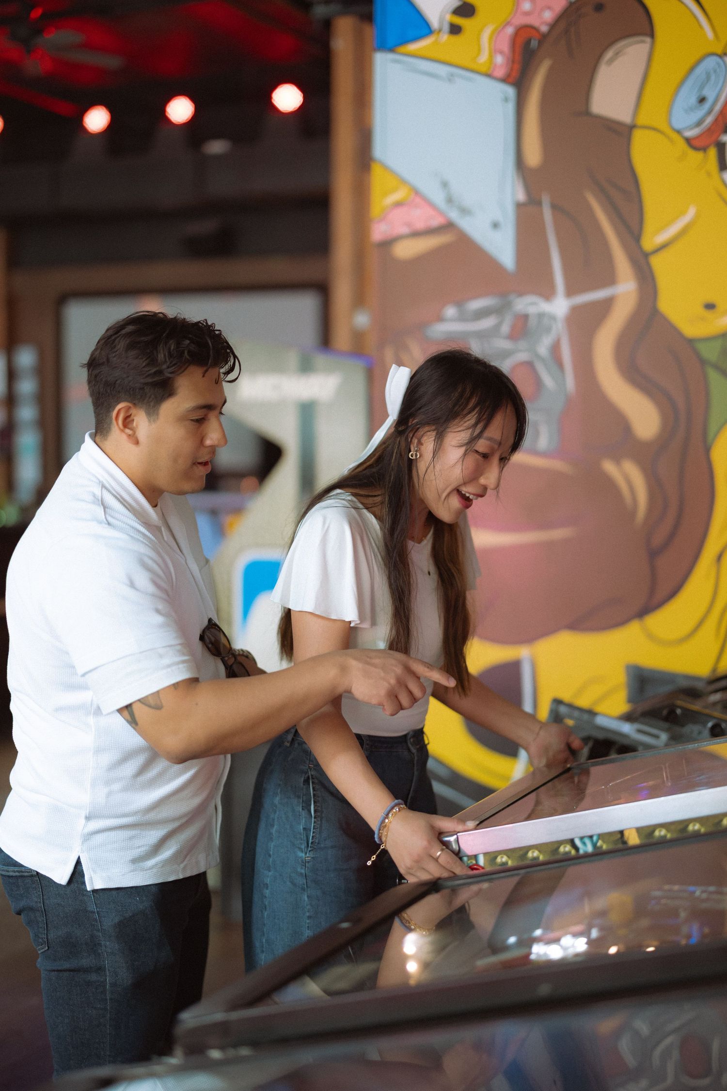 Young couple laughing and pointing at an arcade game in a colorful entertainment venue.