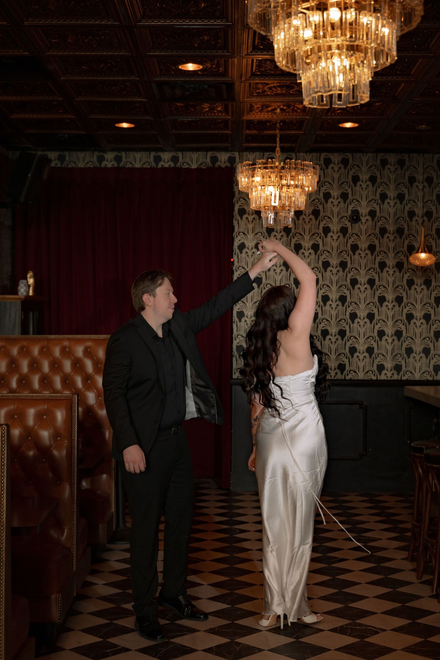 Bride in white satin gown being twirled by groom in black suit under crystal chandeliers in elegant venue.