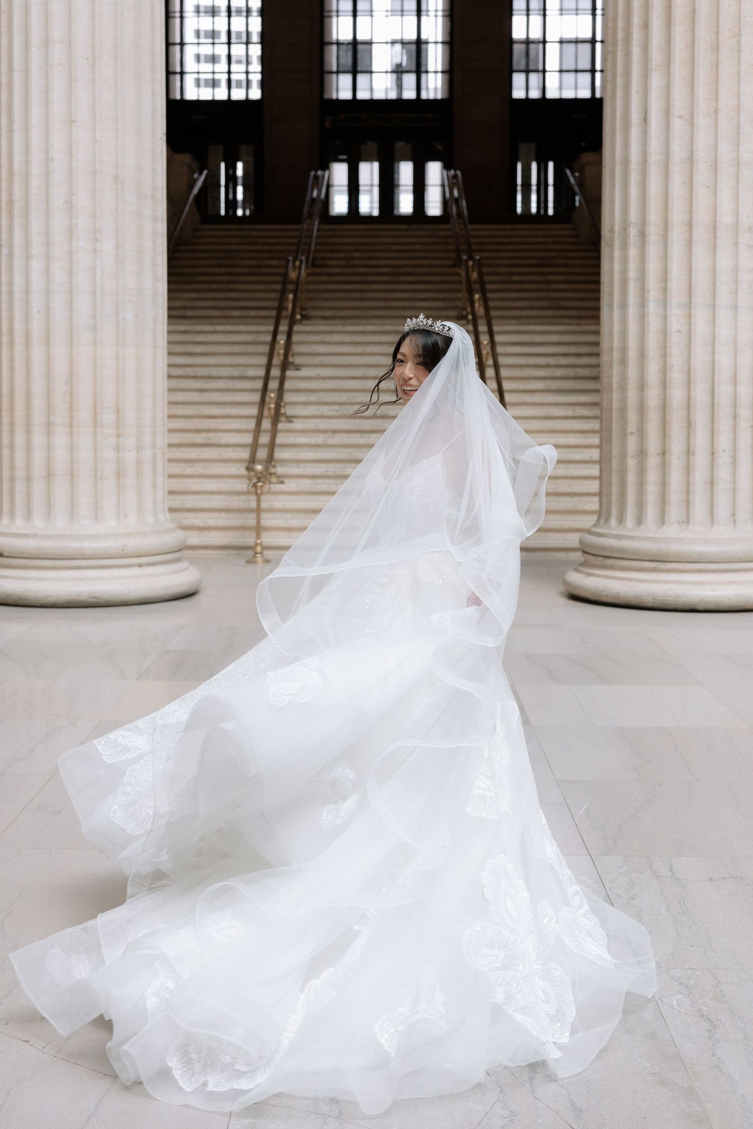 Bride in a flowing white ballgown and tiara with veil billowing between grand marble columns.
