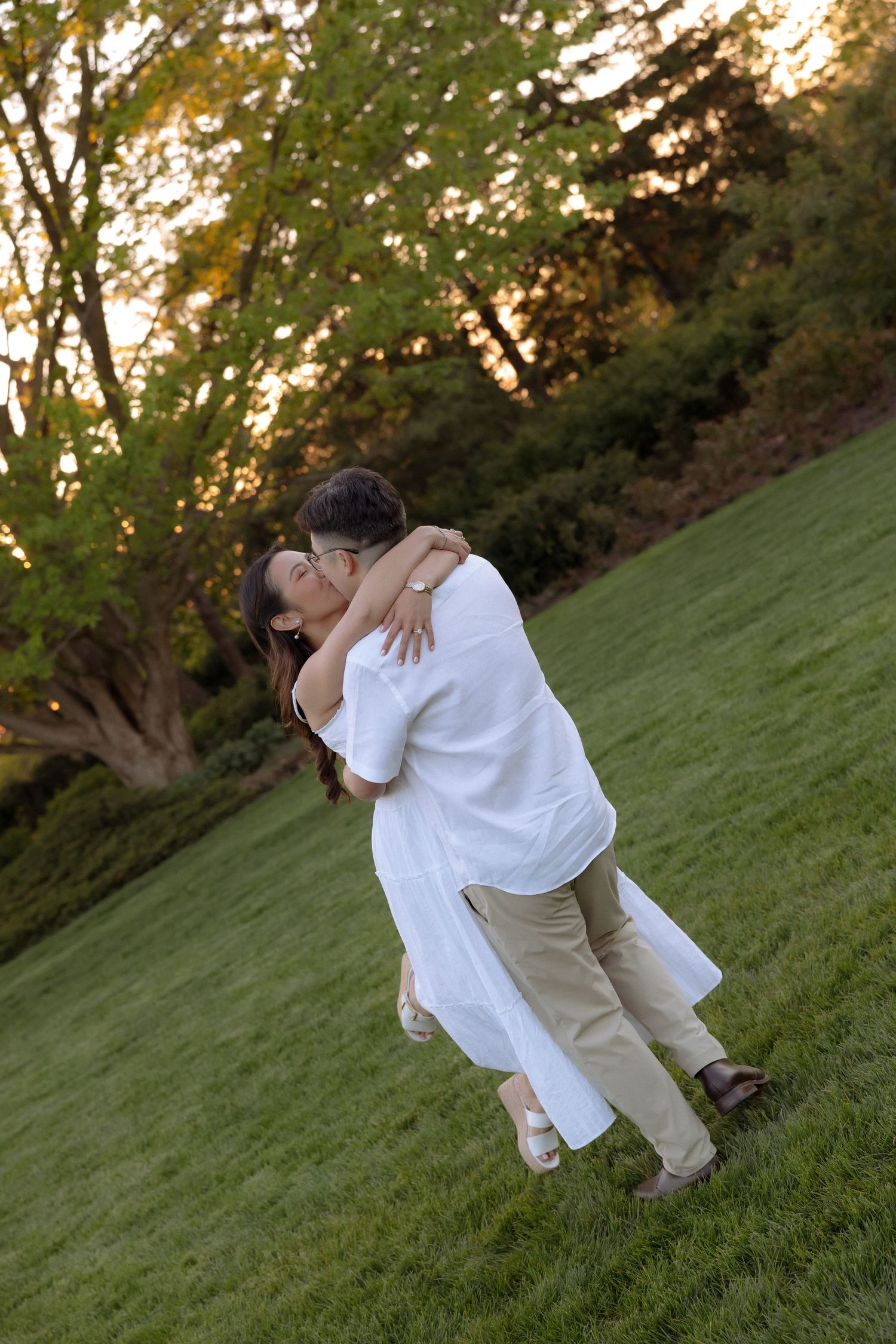 Couple sharing a romantic kiss on a lush green lawn at sunset, surrounded by trees in warm golden light.