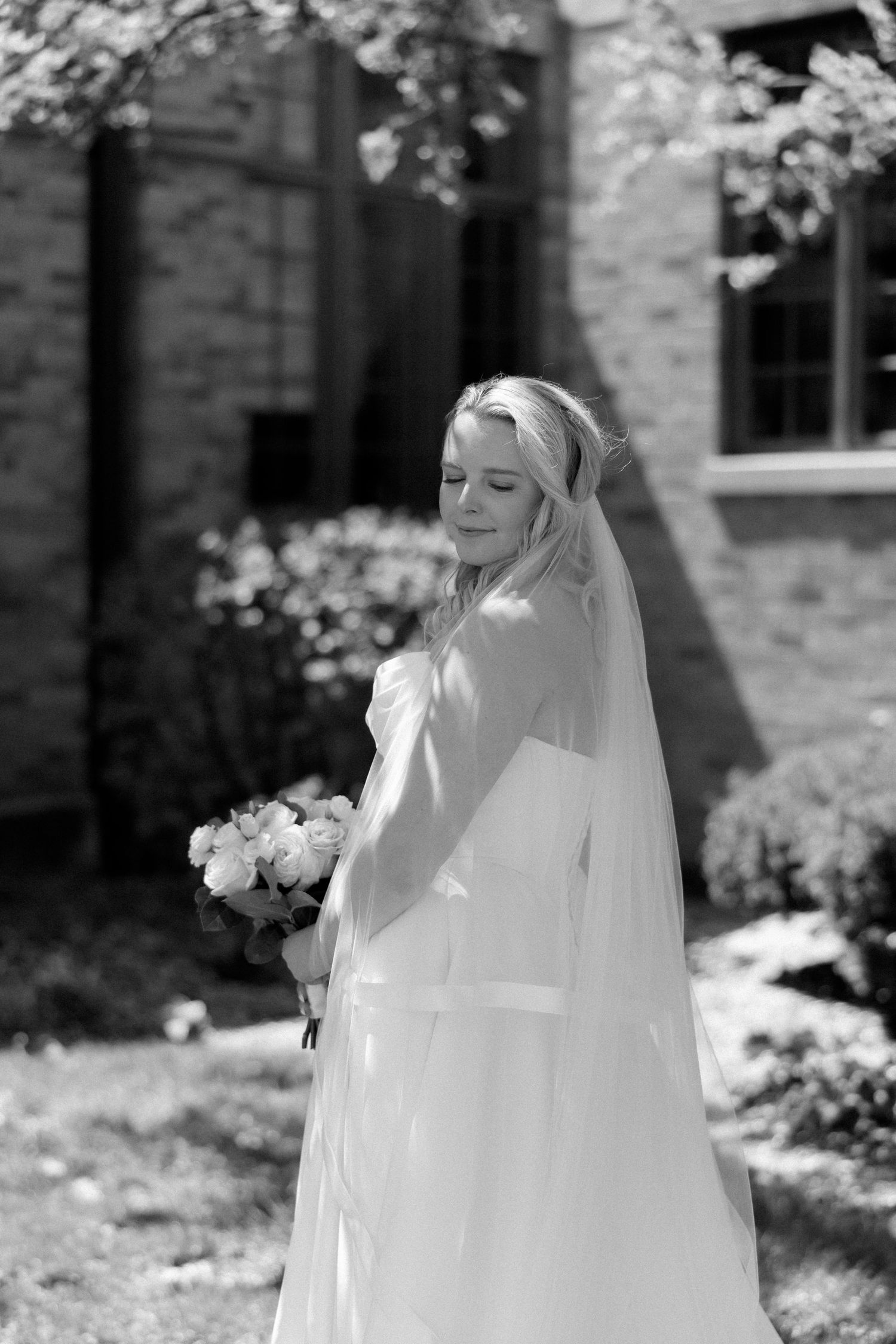 Black and white portrait of a bride in a white gown and veil holding a bouquet outdoors near a brick building.