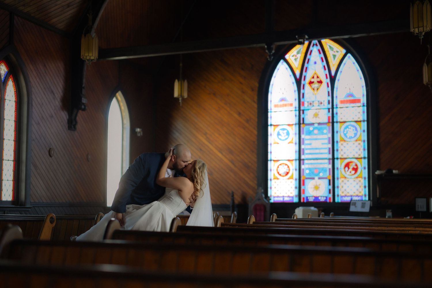 Bride and groom share a kiss in a wooden church pew with colorful stained glass windows in the background.
