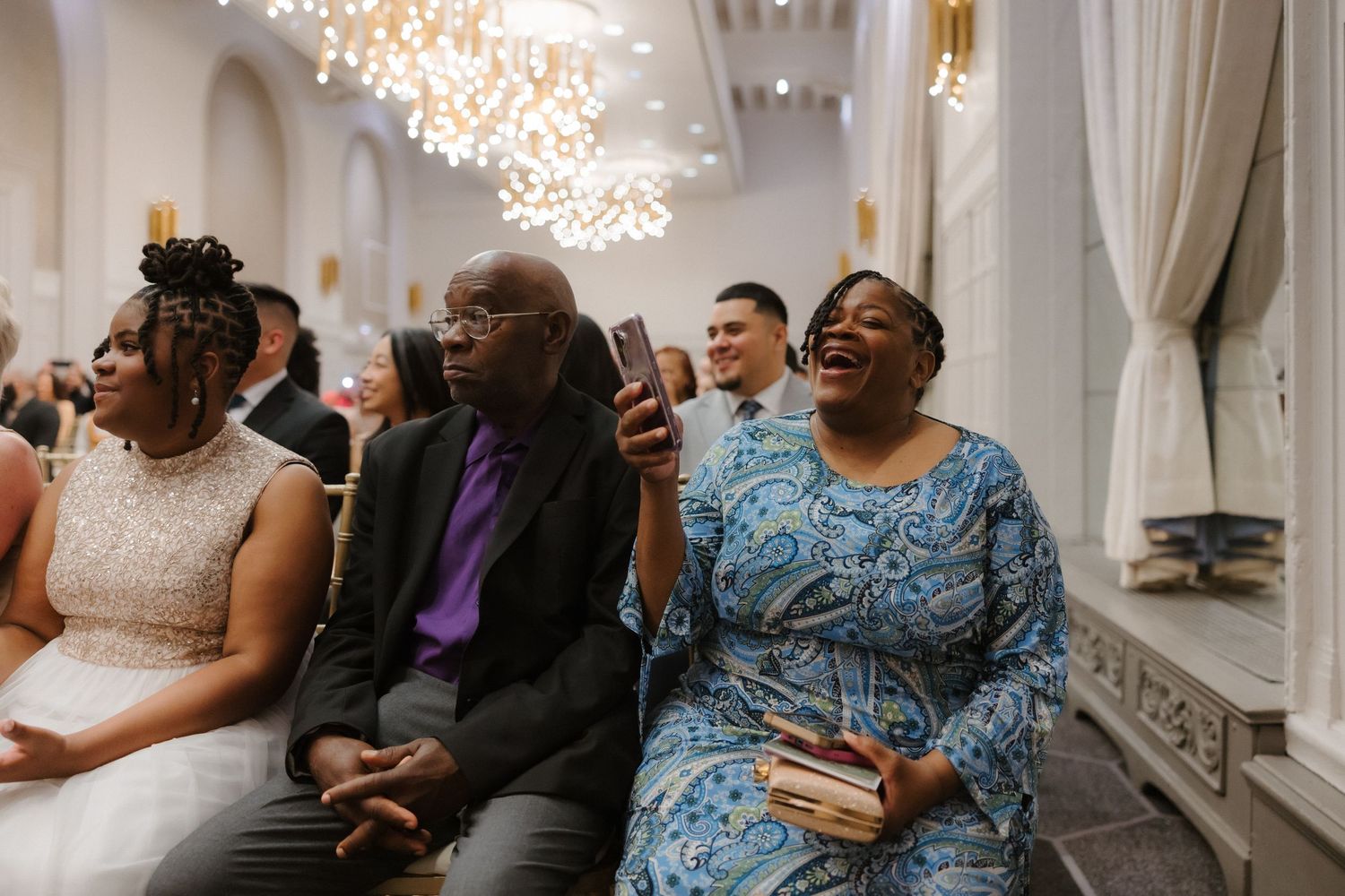 Joyful wedding guests laughing and smiling during a ceremony in an elegant venue with chandelier lighting.