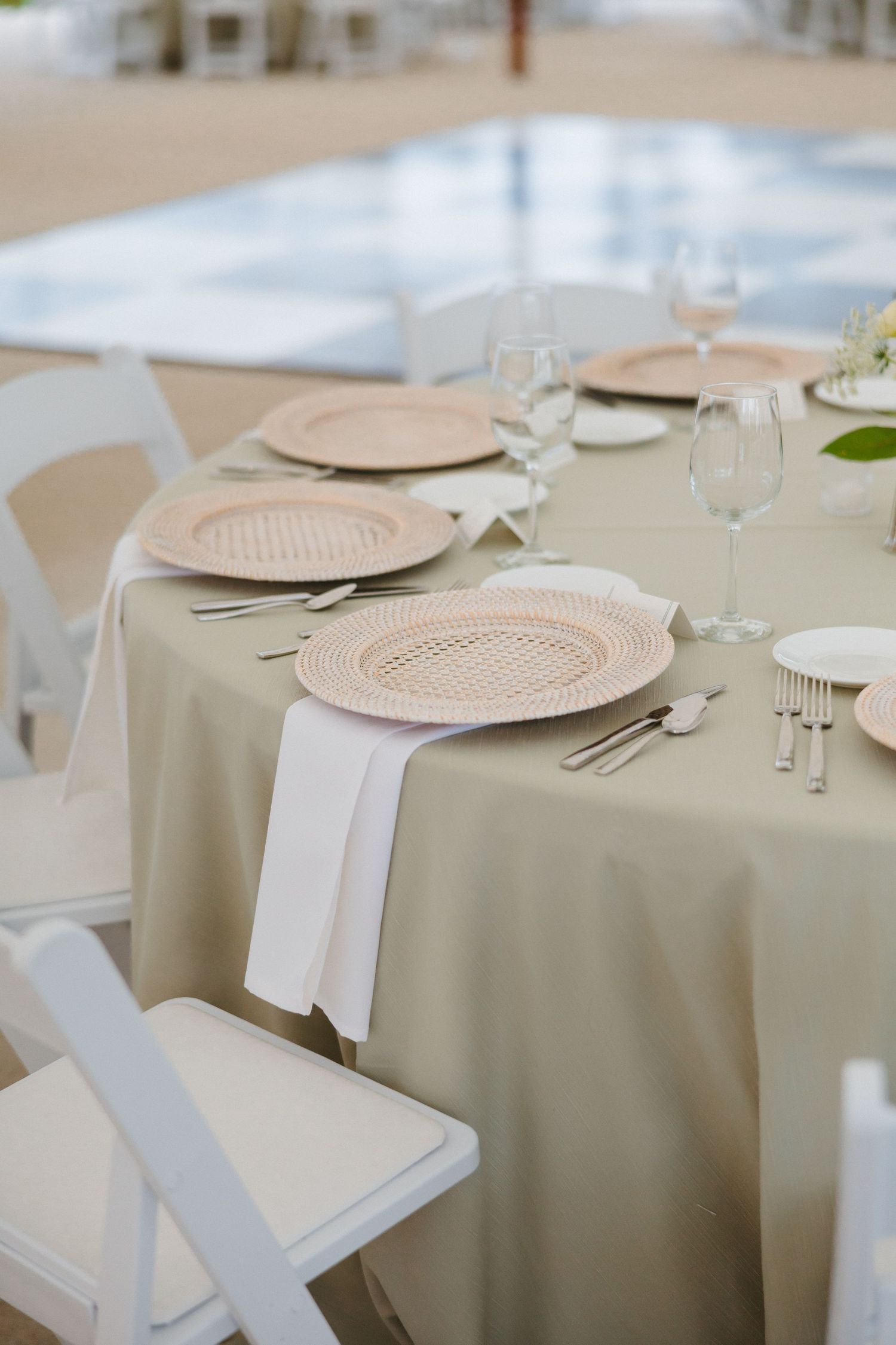 Round wedding reception table with woven charger plates, sage green linen, and white folding chairs.