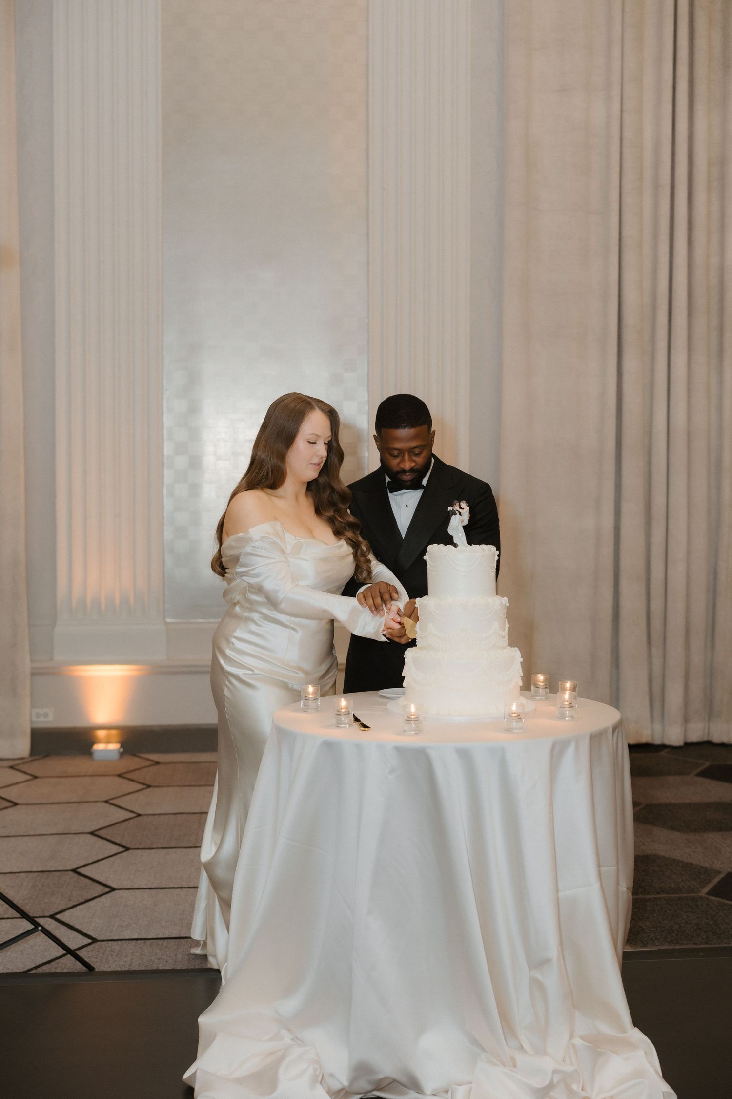 Bride and groom cutting a white tiered wedding cake on a candlelit table in an elegant ballroom.