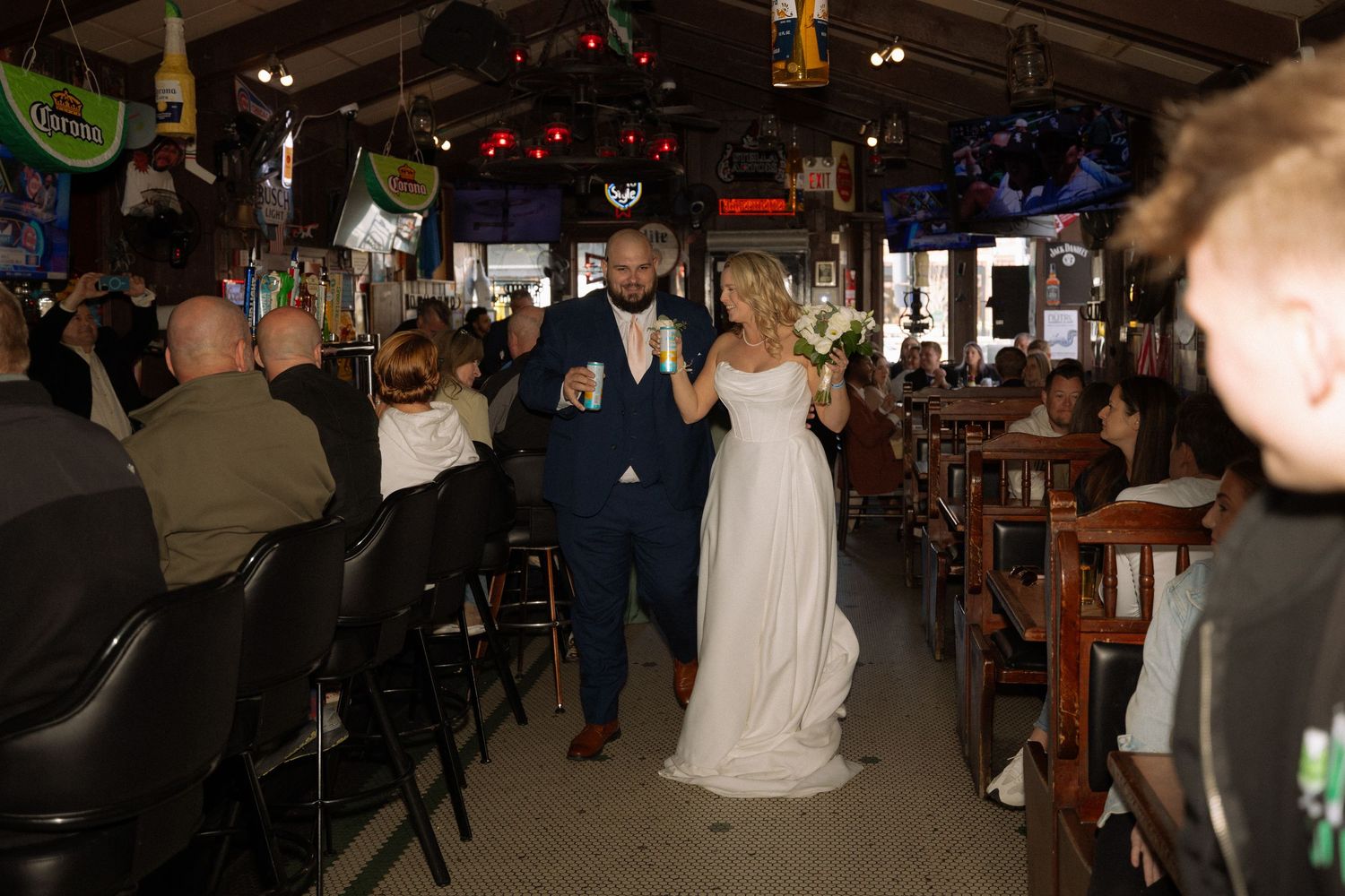 Bride and groom walk down the aisle of a rustic bar venue, smiling and holding drinks during their wedding reception.