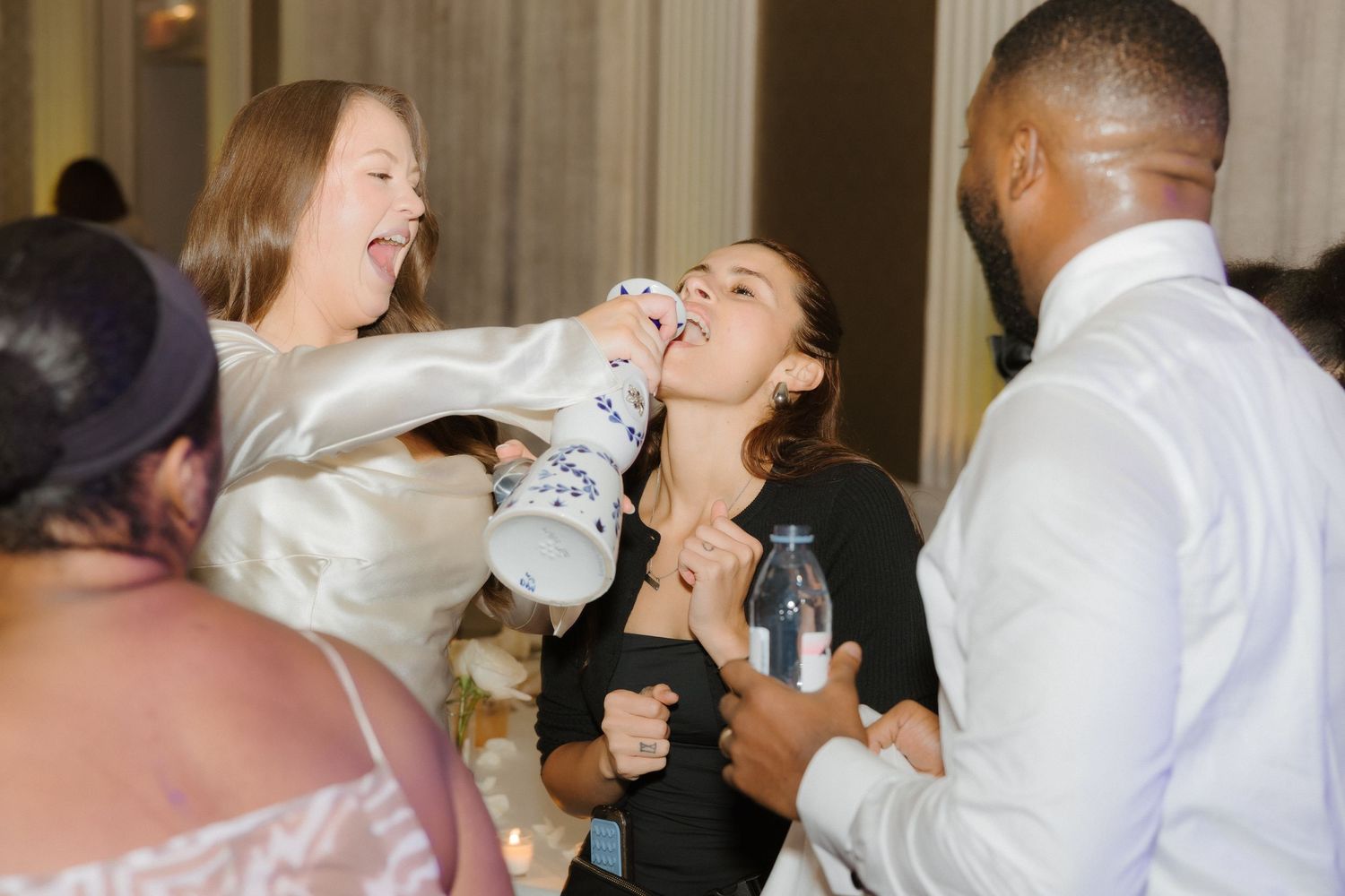 Two women laughing and dancing joyfully at a social gathering or wedding reception with other guests nearby.
