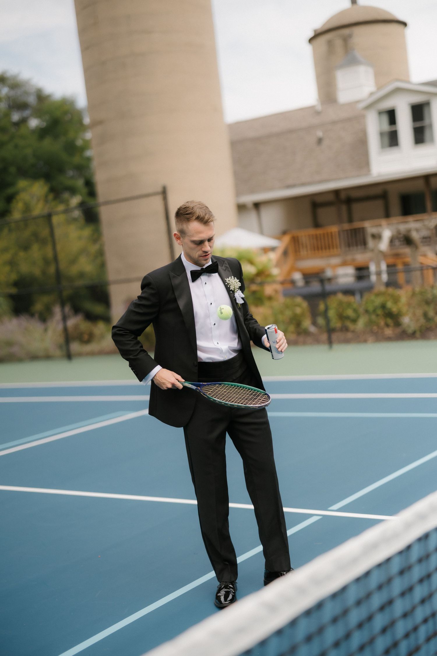 Dapper groom in black tuxedo with boutonniere holds tennis racket on blue court before wedding.