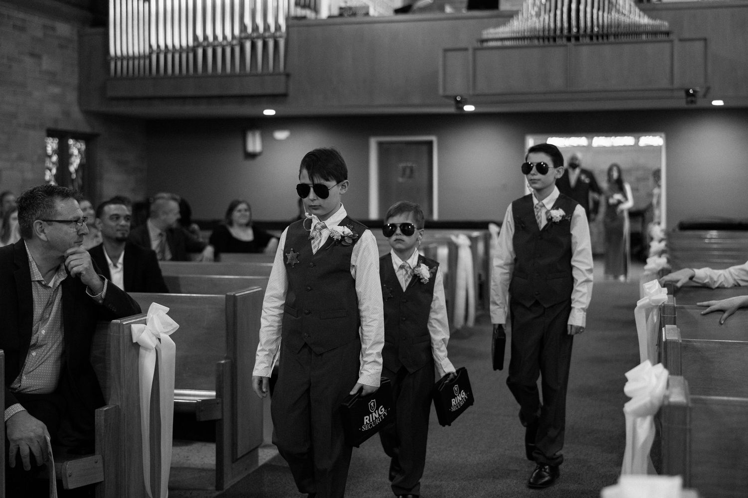 Three young boys in vests and sunglasses walk down a church aisle during a wedding ceremony.