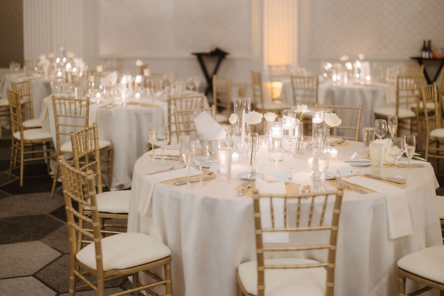 Elegant wedding reception hall with gold Chiavari chairs, white round tables, and glowing candle centerpieces.
