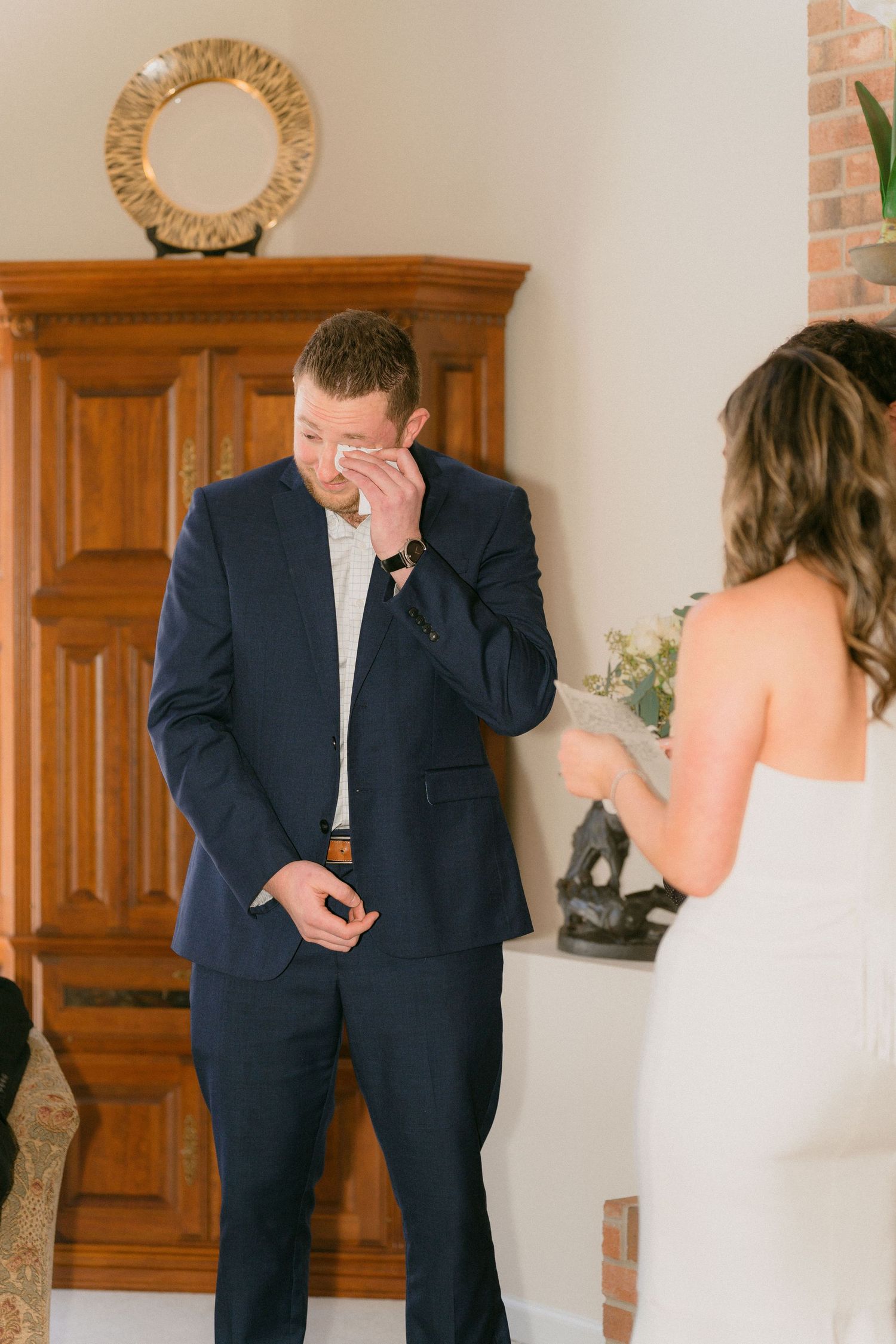 Emotional groom wiping tears in navy suit during first look at wedding.