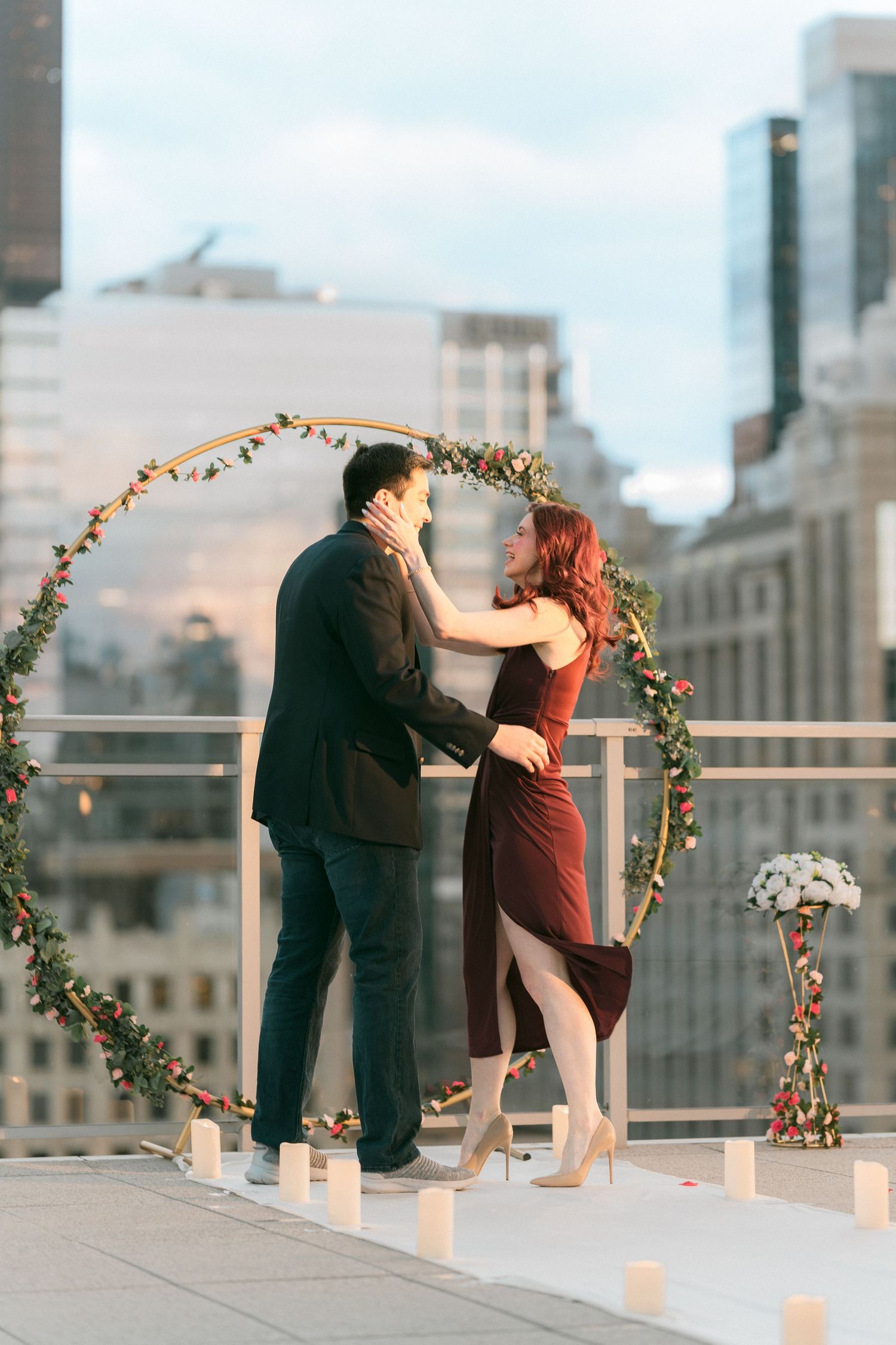 Couple embracing under a floral hoop arch on a rooftop with a city skyline at dusk.