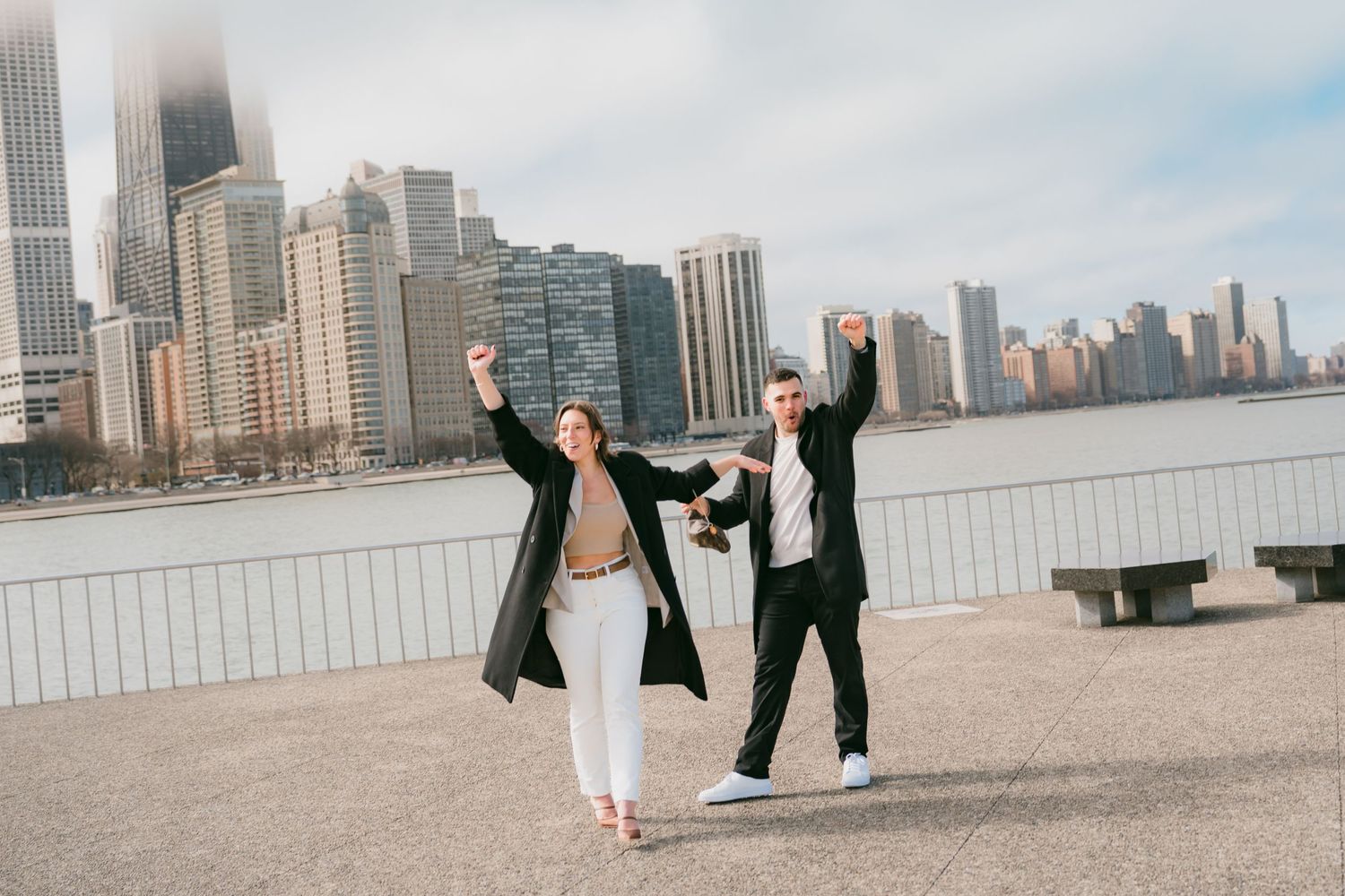 Excited newly engaged couple celebrating with raised arms along Chicago lakefront waterfront promenade.