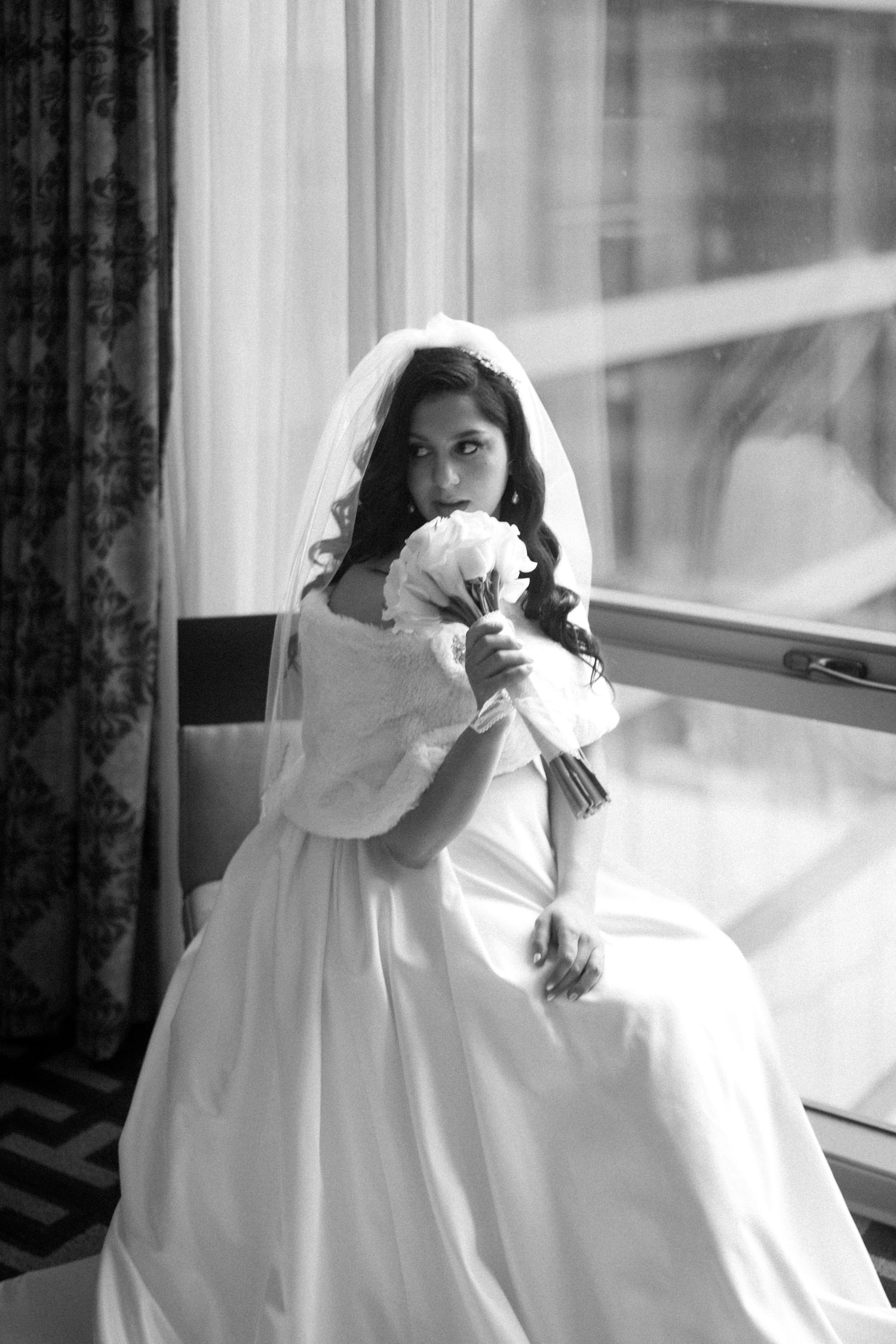 Black and white portrait of a veiled bride in a full gown holding a bouquet by a window.