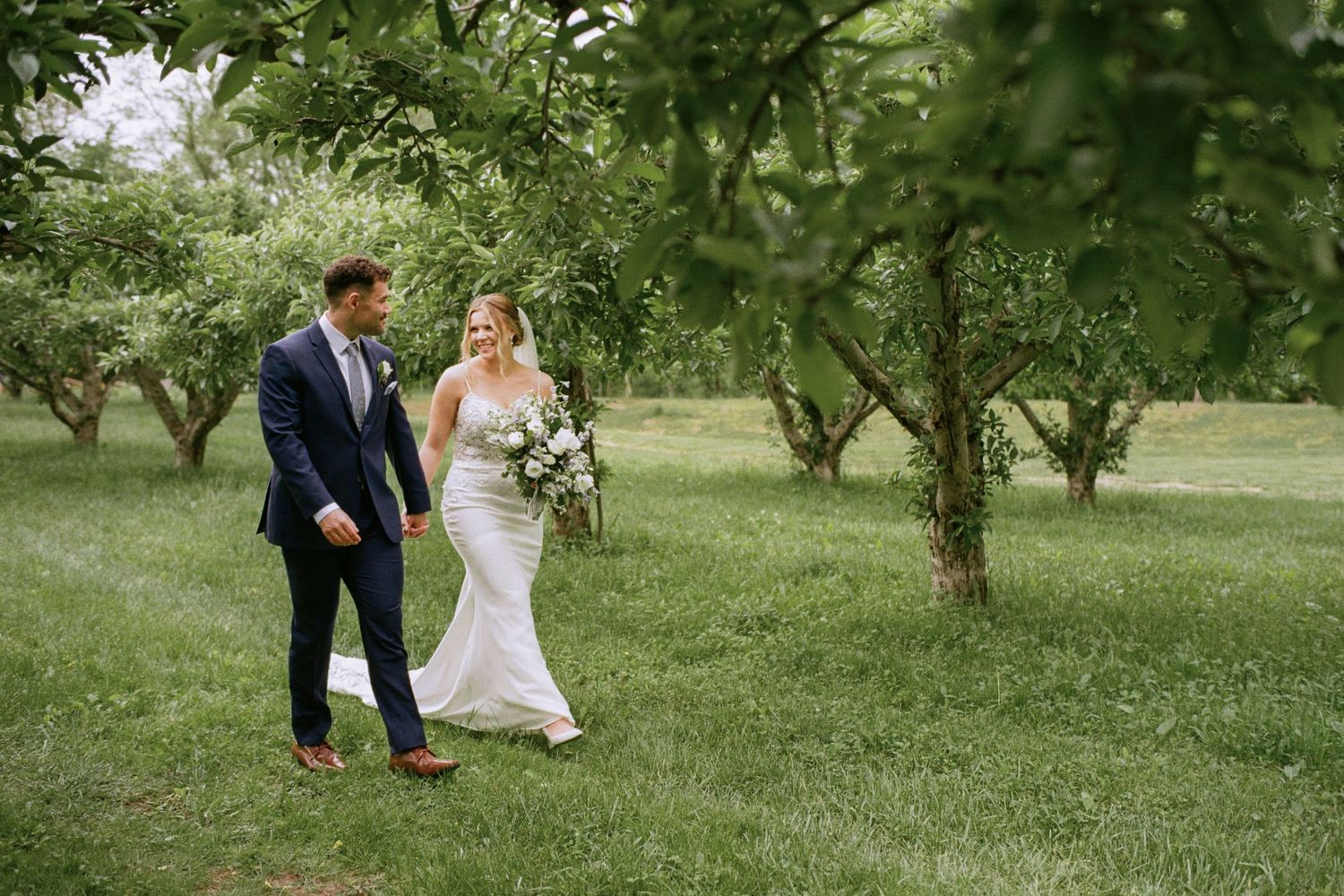 Bride and groom walking hand in hand through a lush green orchard on their wedding day.
