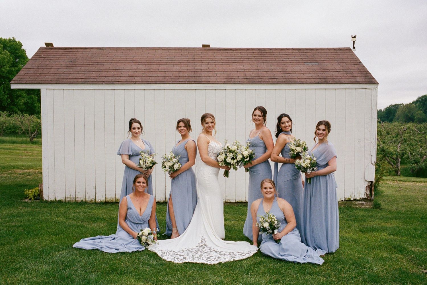 Bride in lace gown poses with six bridesmaids in dusty blue dresses holding bouquets before a white barn.