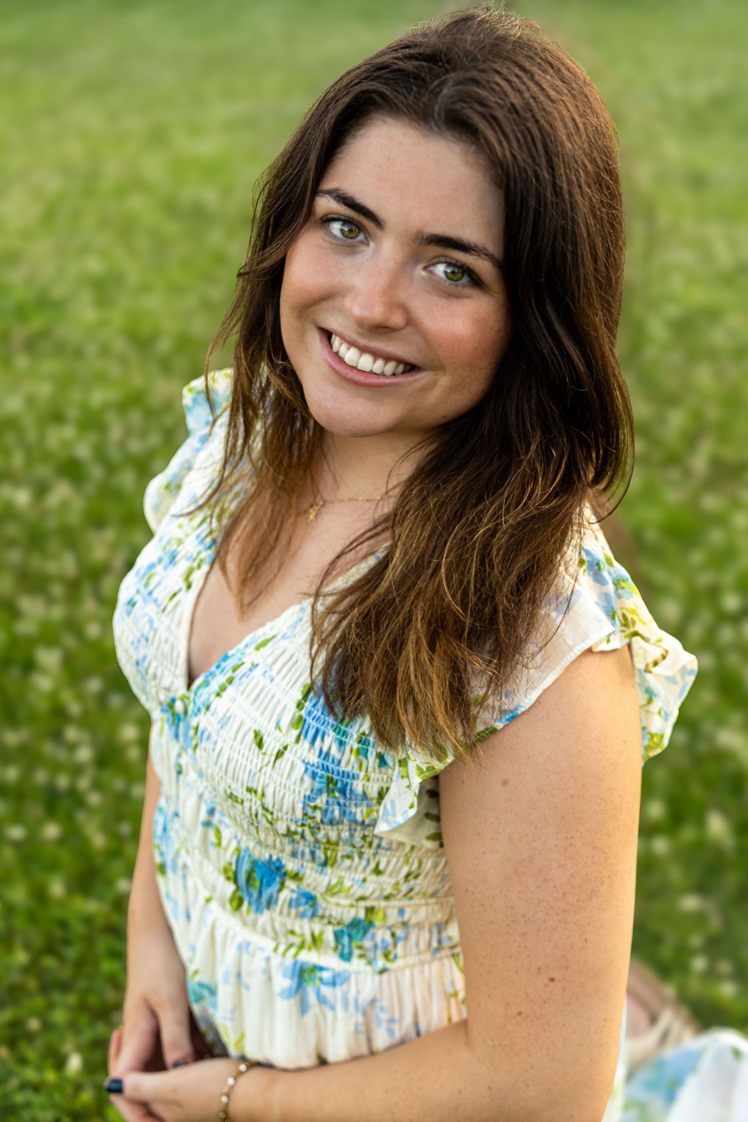 Smiling young woman with brown hair wearing a floral dress, sitting outdoors in a green field on a sunny day.