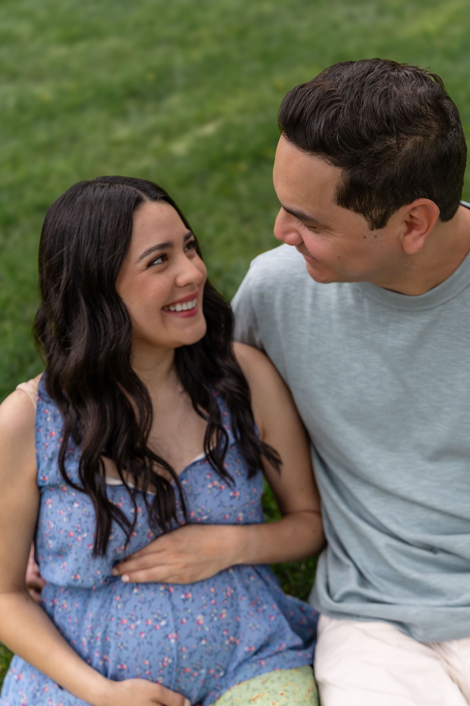 Happy pregnant couple sitting on grass, smiling at each other, woman in blue floral dress cradling baby bump.