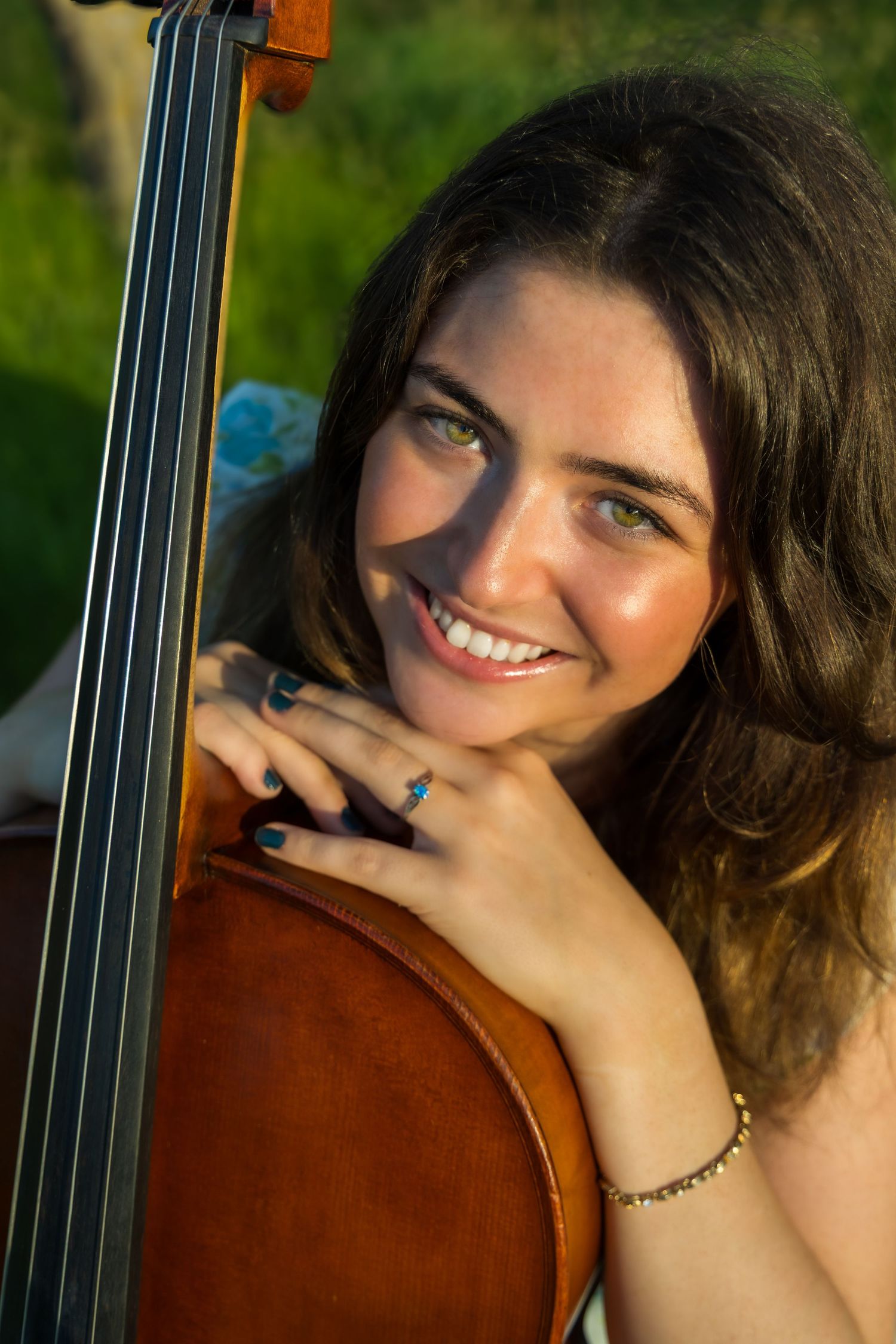 Smiling young woman with green eyes resting her chin on a cello outdoors in warm sunlight.