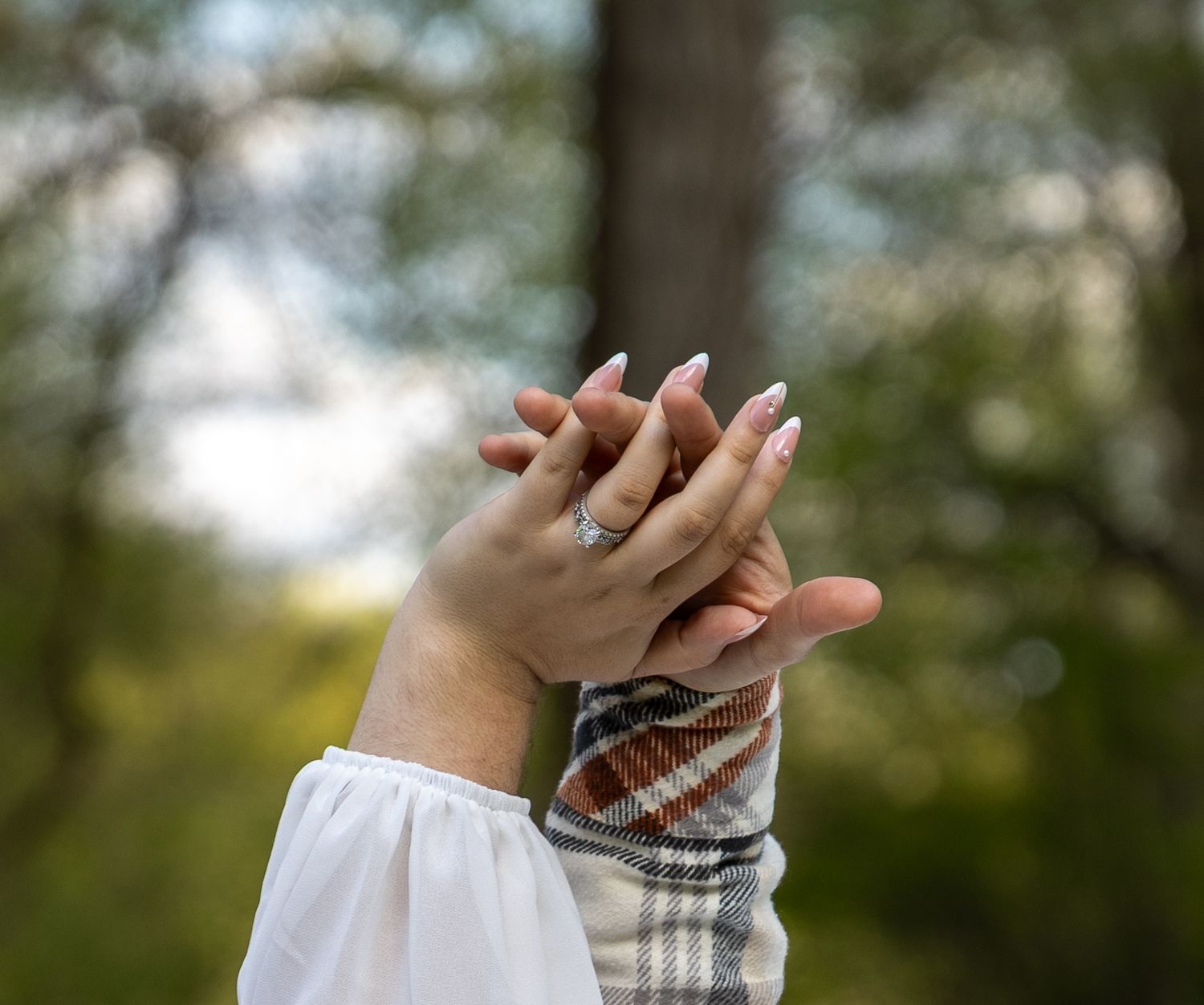 Two hands intertwined outdoors, one wearing a diamond engagement ring, with a blurred green forest background.