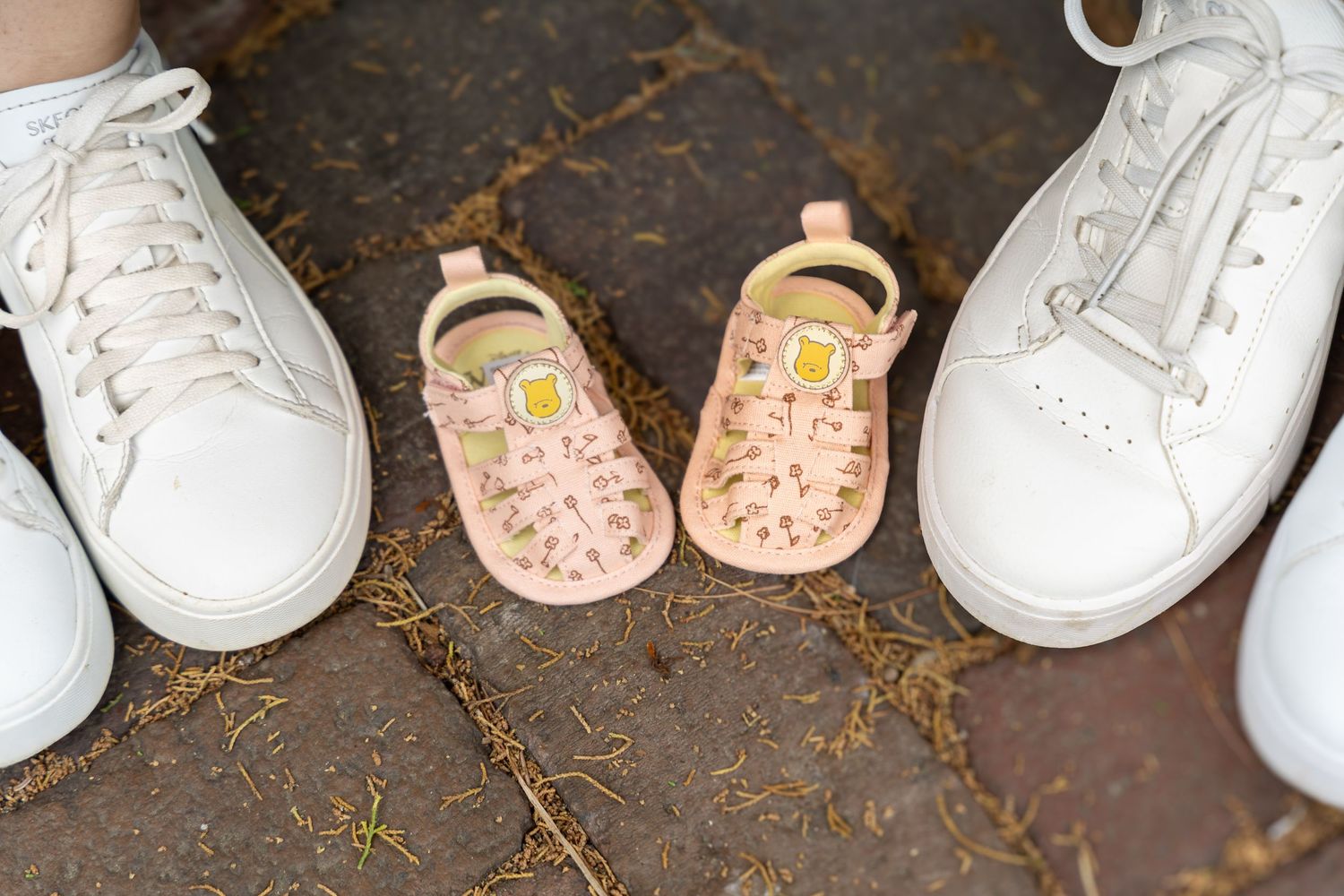 Two white adult sneakers flank tiny pink baby shoes with gold accents on a textured ground.