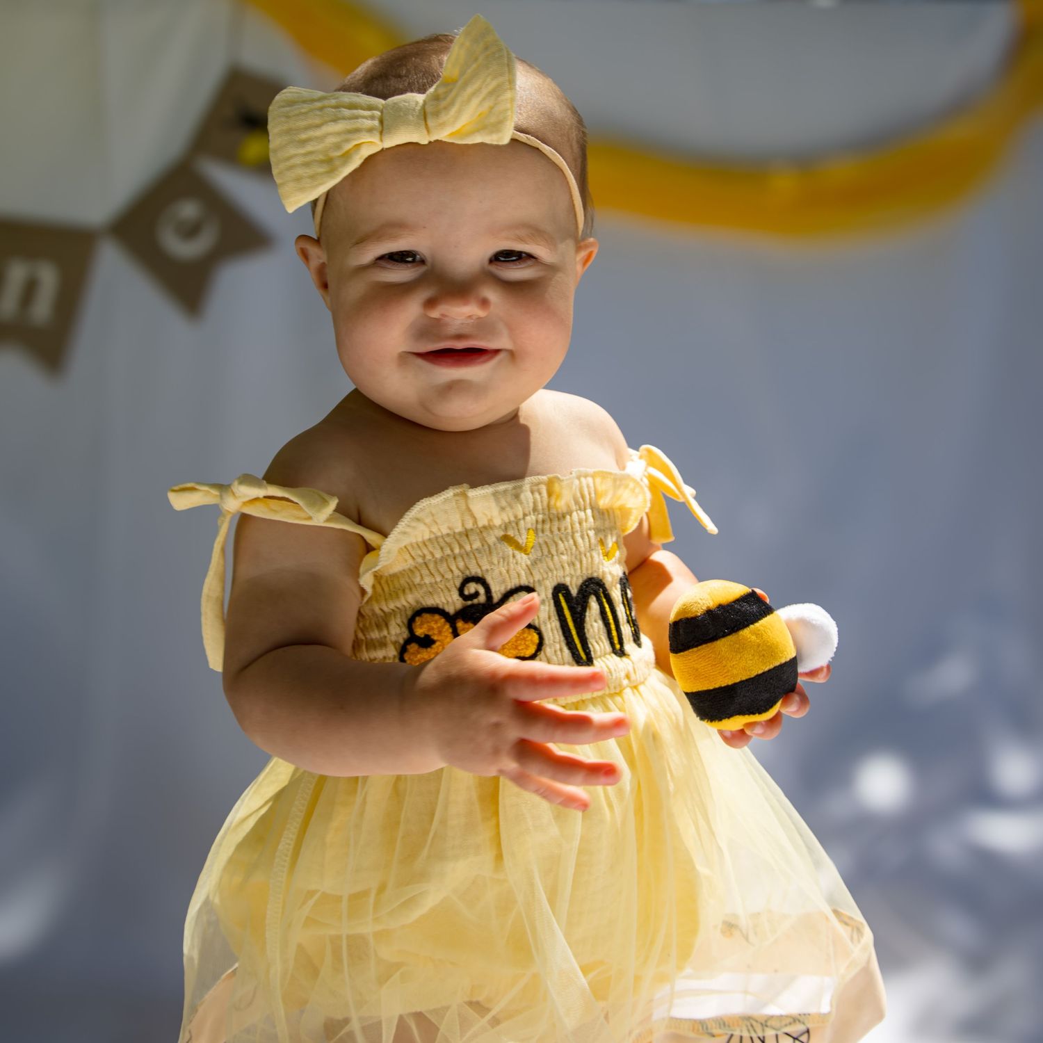 Baby girl in yellow tutu dress and bow headband holds a bumblebee toy at a bee-themed first birthday party.