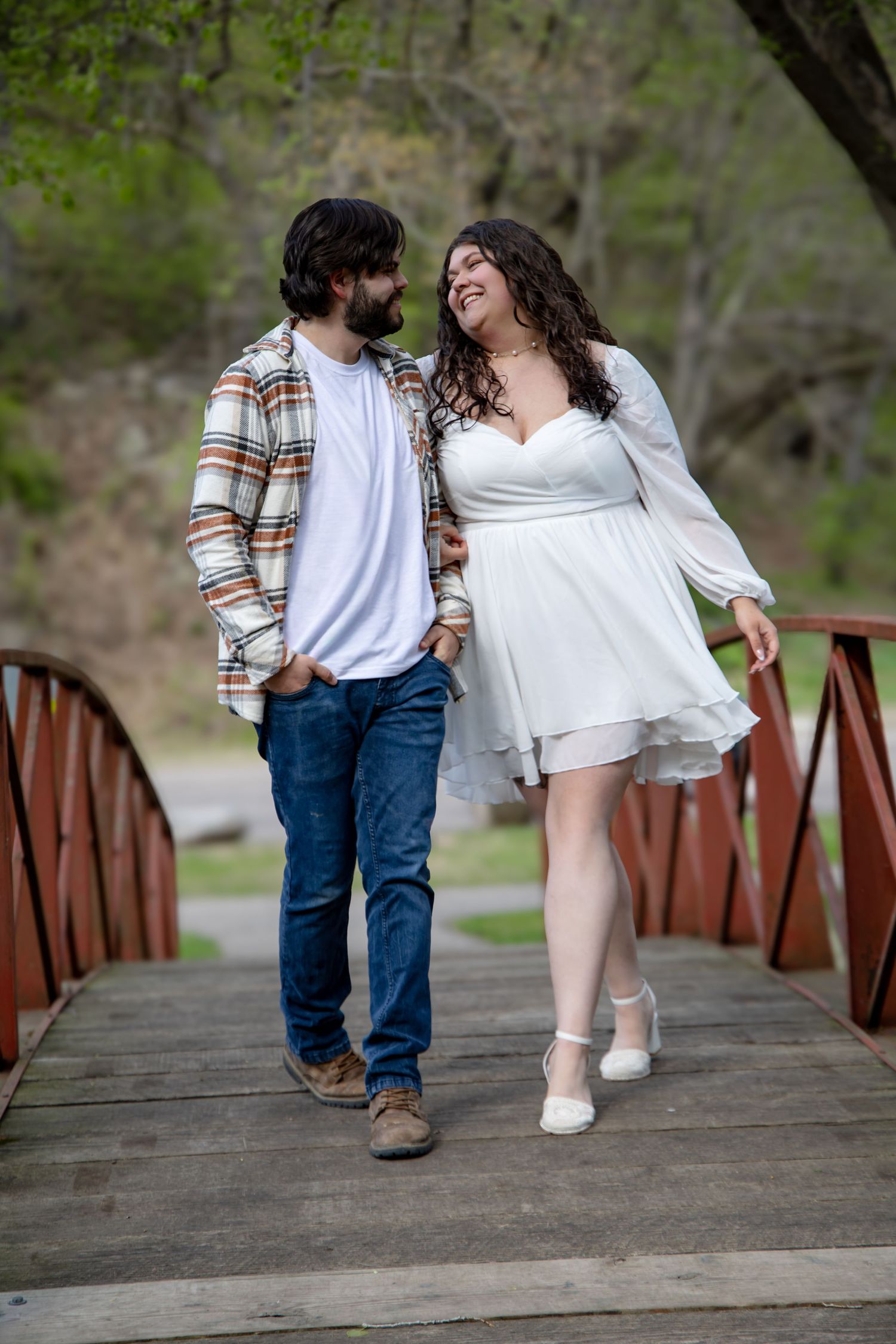Couple laughing and walking together on a wooden bridge in a lush green park setting.