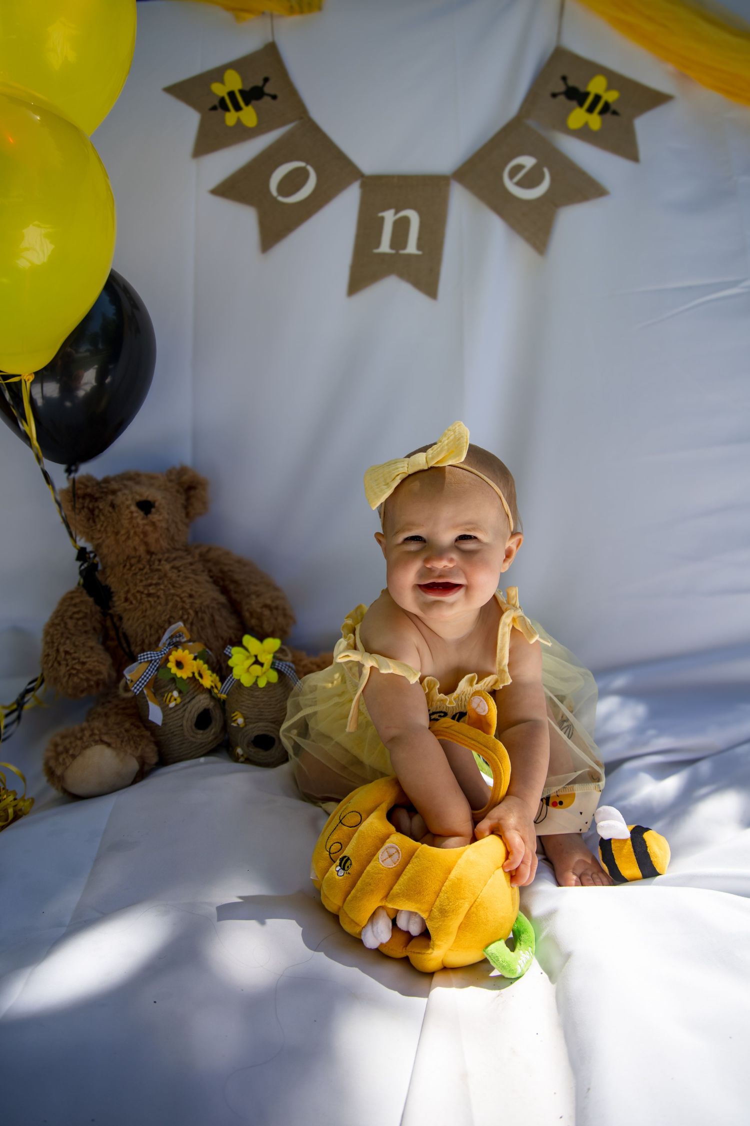 Smiling baby girl in yellow tutu sits at bee-themed first birthday party setup with stuffed animals and ONE banner.