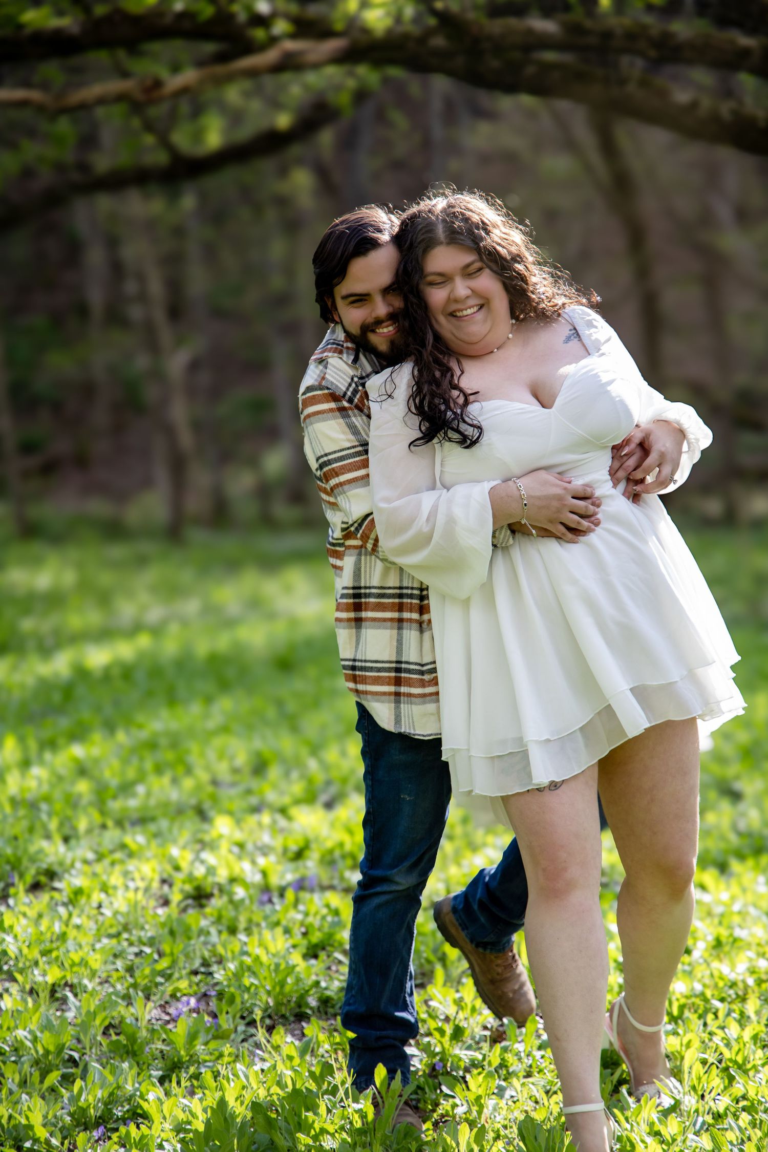 Couple embracing outdoors in green field, woman in white dress, man in plaid shirt, smiling joyfully among spring foliage.
