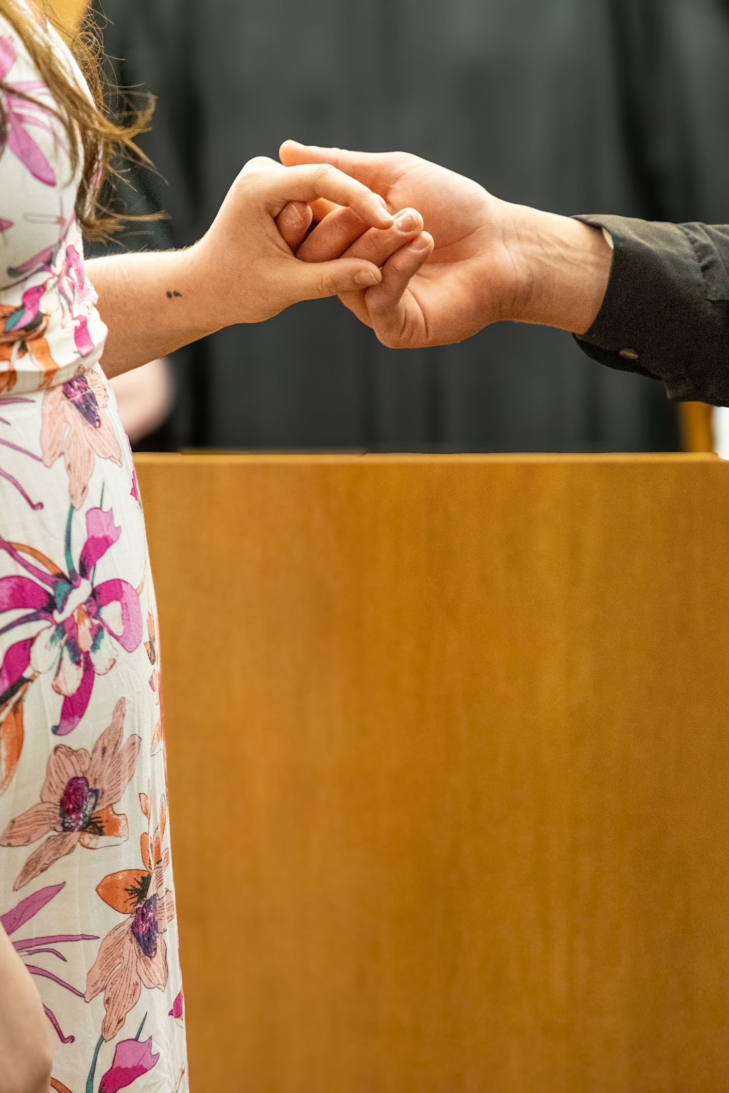Couple gently holding hands in front of a wooden podium, woman wearing a floral dress.