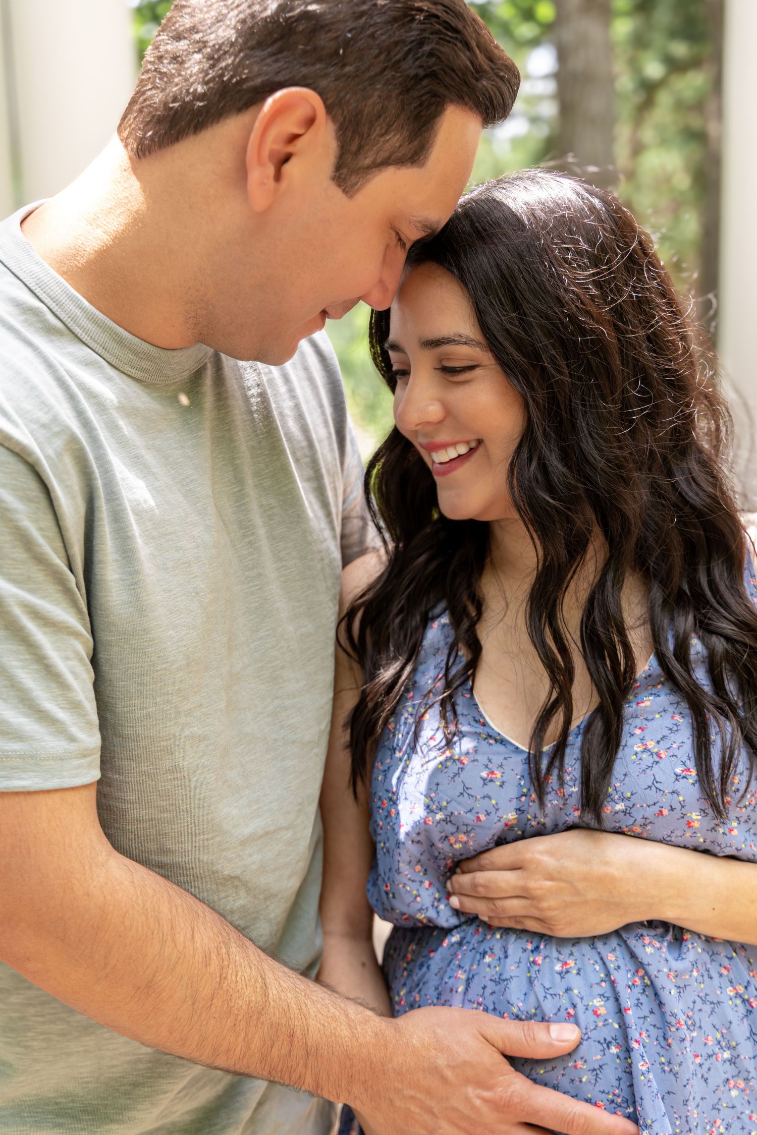 A happy couple shares a tender moment, with the pregnant woman in a blue floral dress cradling her baby bump.