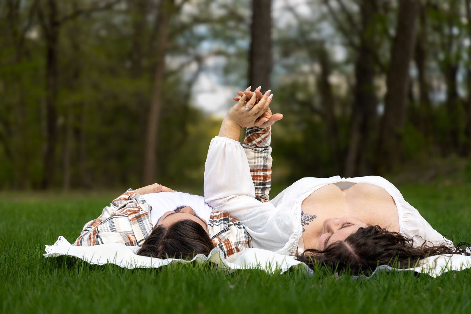 Two women lying on green grass in a park, wearing white and plaid outfits, relaxing outdoors on a sunny day.