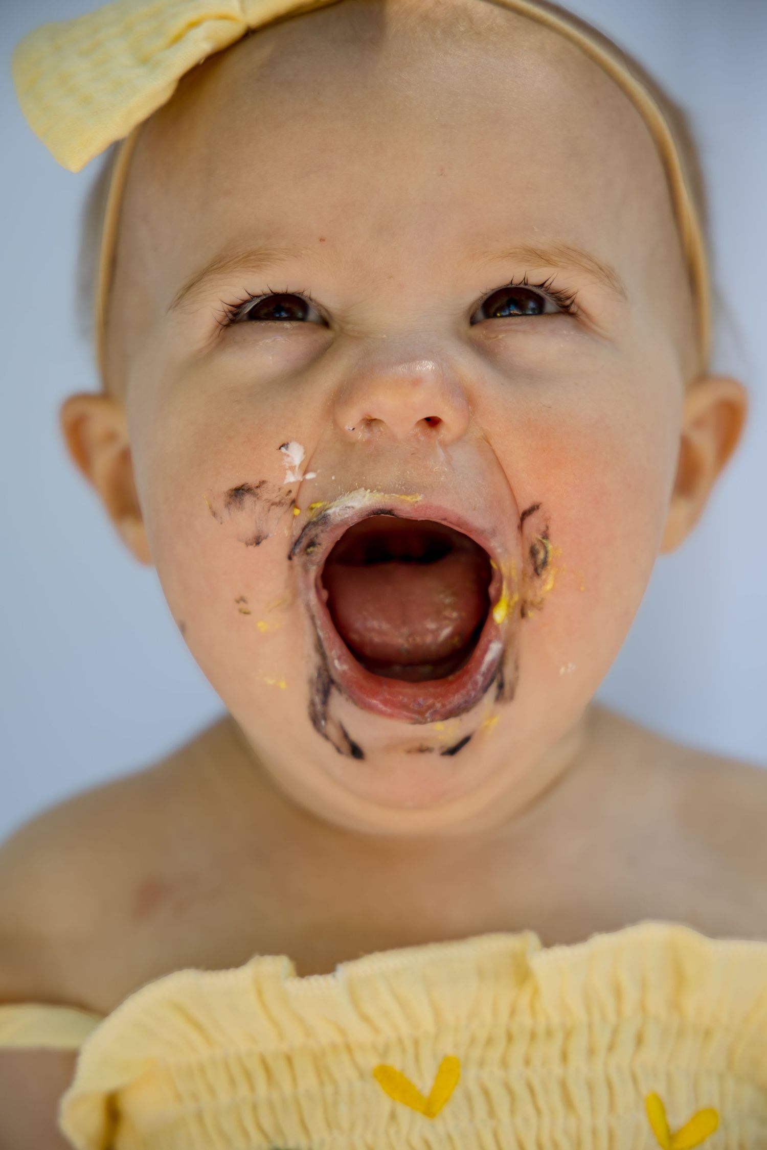 Baby girl with cake smeared on her face, mouth wide open, wearing yellow bow and outfit during first birthday cake smash.