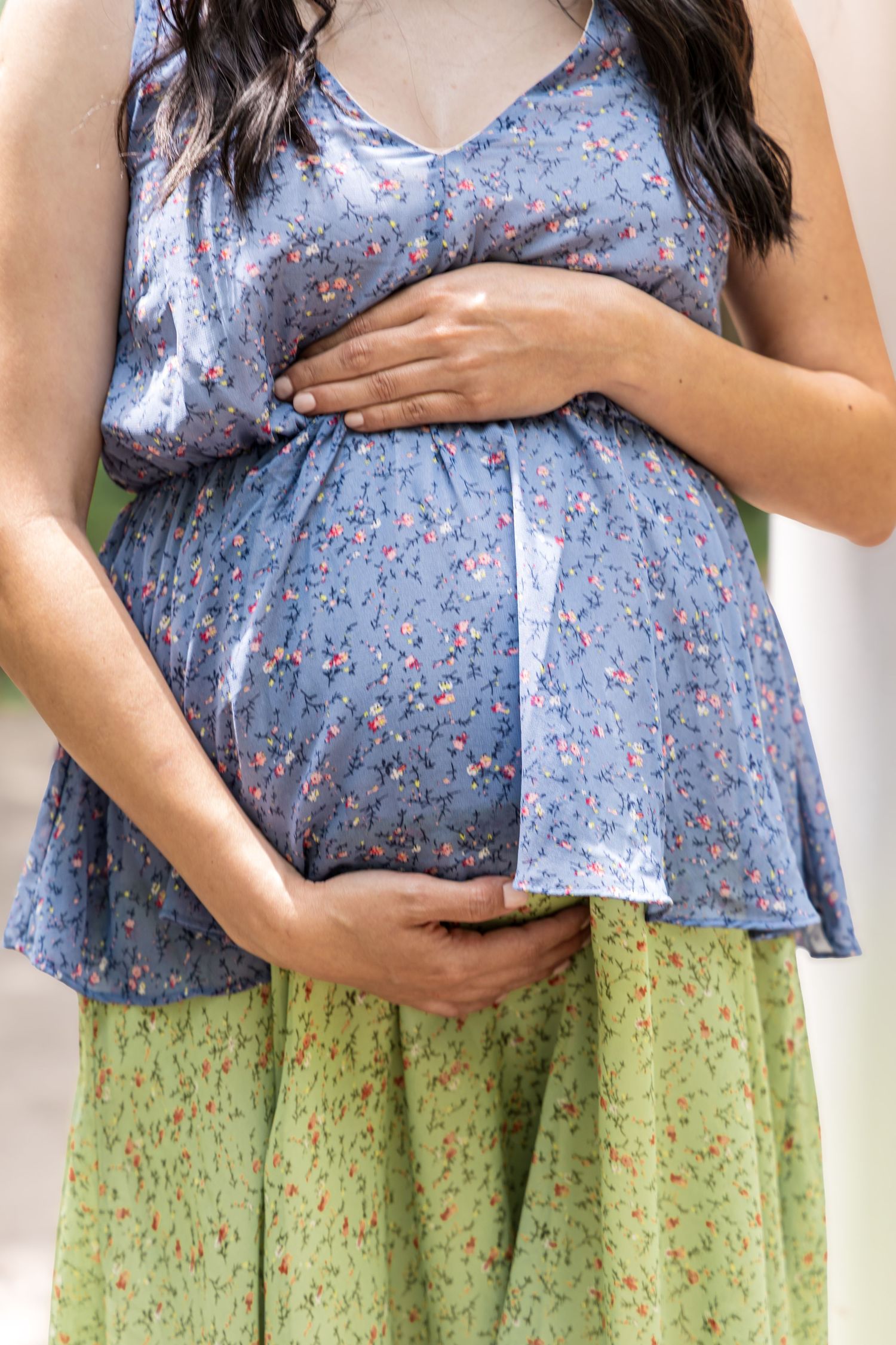 Pregnant woman in blue floral top and green skirt cradling her baby bump with both hands outdoors.