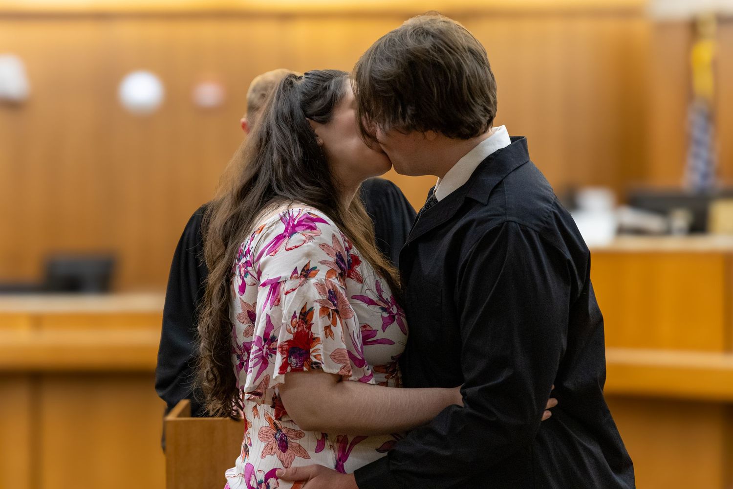 Couple sharing a kiss in a courtroom setting, both wearing black robes during a ceremony.