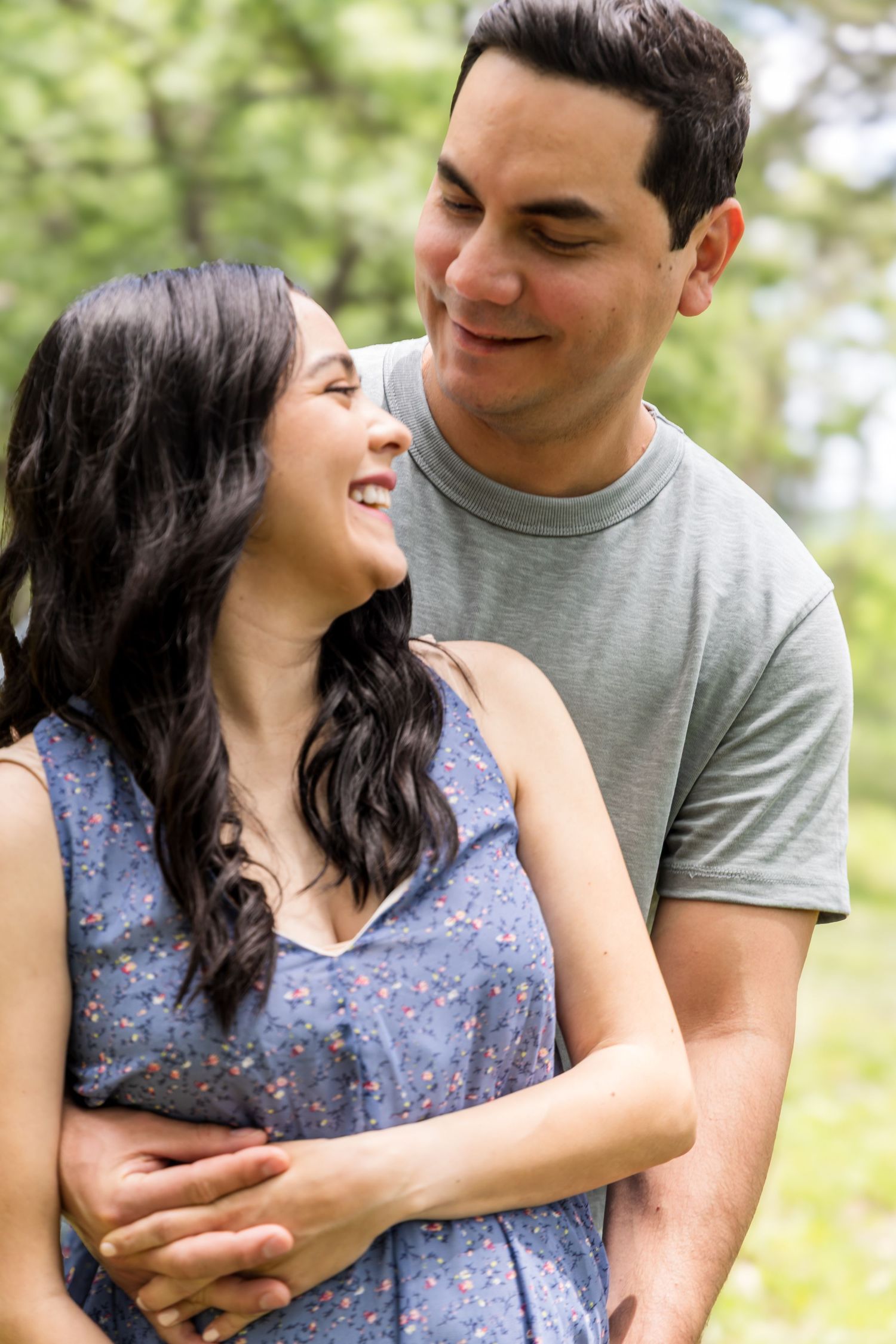 Happy couple embracing outdoors, woman in blue floral dress smiling up at man in gray t-shirt in green park setting.