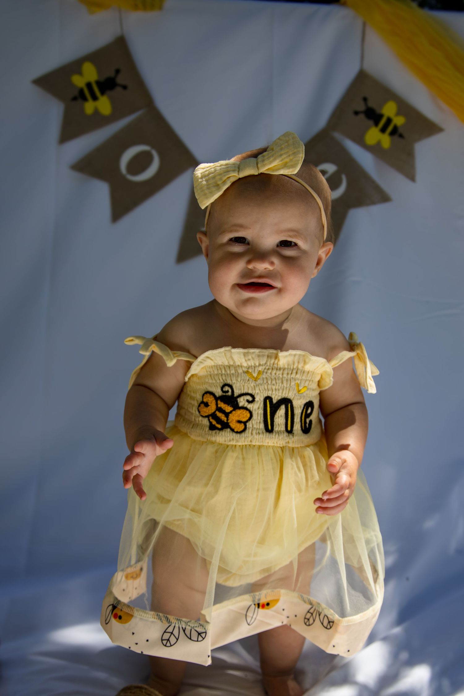 Adorable baby girl in yellow tulle bee dress stands at first birthday party with bee banner decoration behind her.