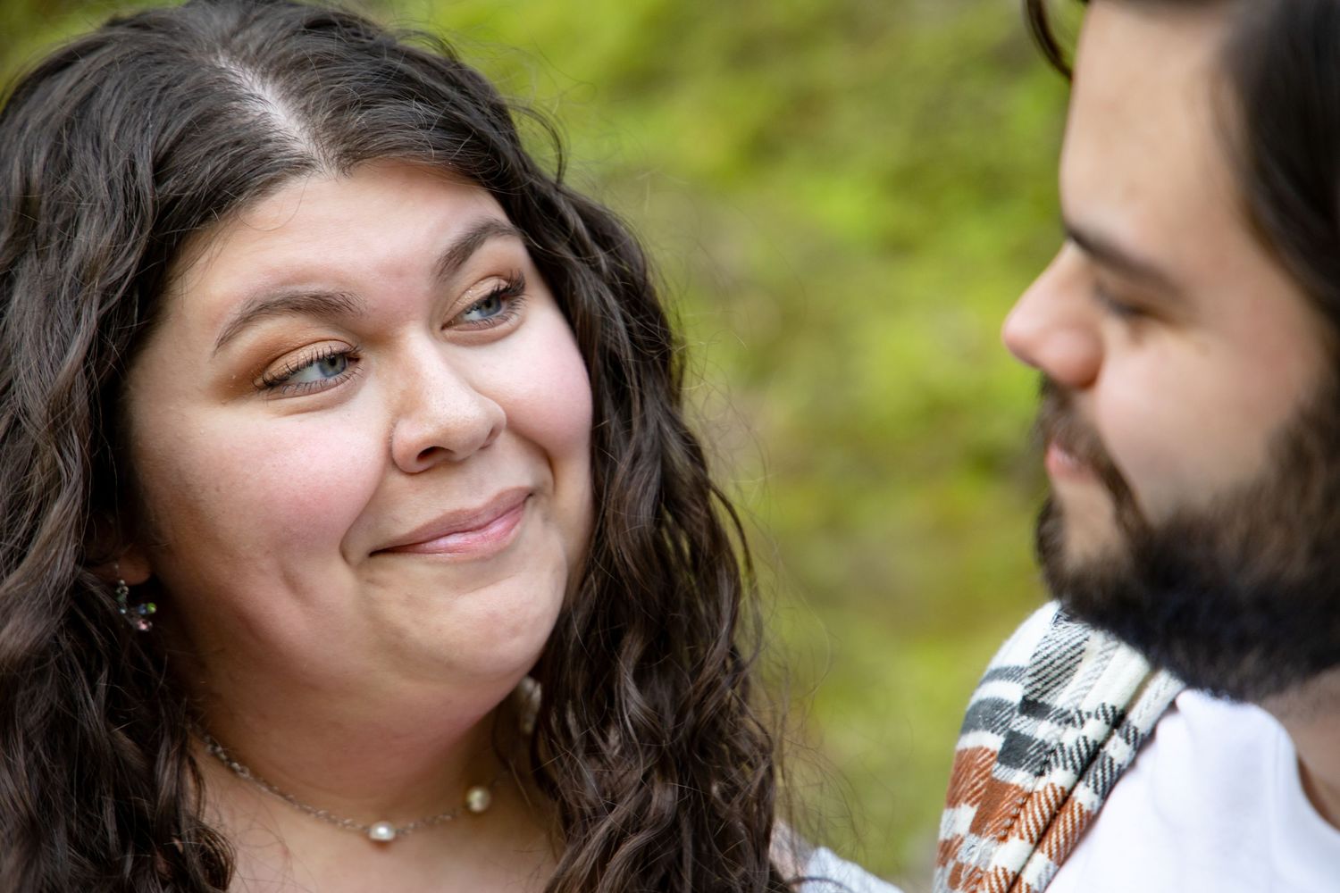 Woman with curly hair smiling at bearded man during outdoor engagement photo session.