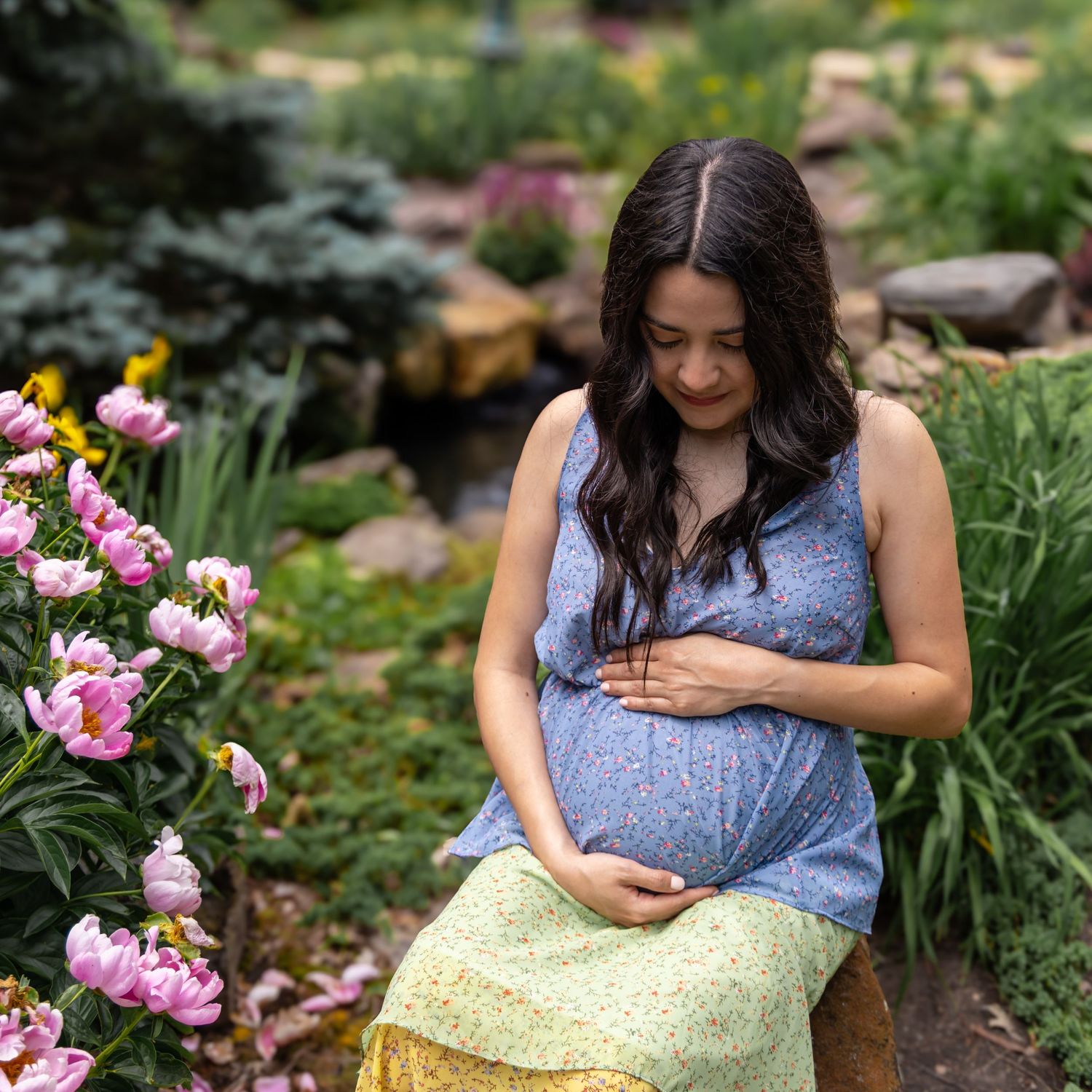 Pregnant woman in blue floral top sits in lush garden with pink peonies, cradling her baby bump.