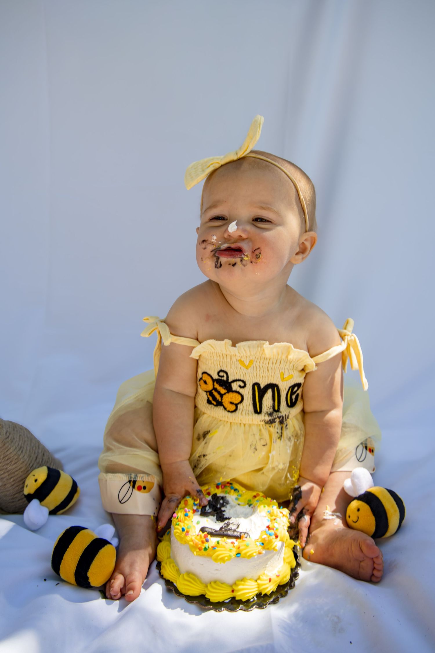 Baby girl in bee costume smashes first birthday cake, face covered in frosting, sitting on white background.