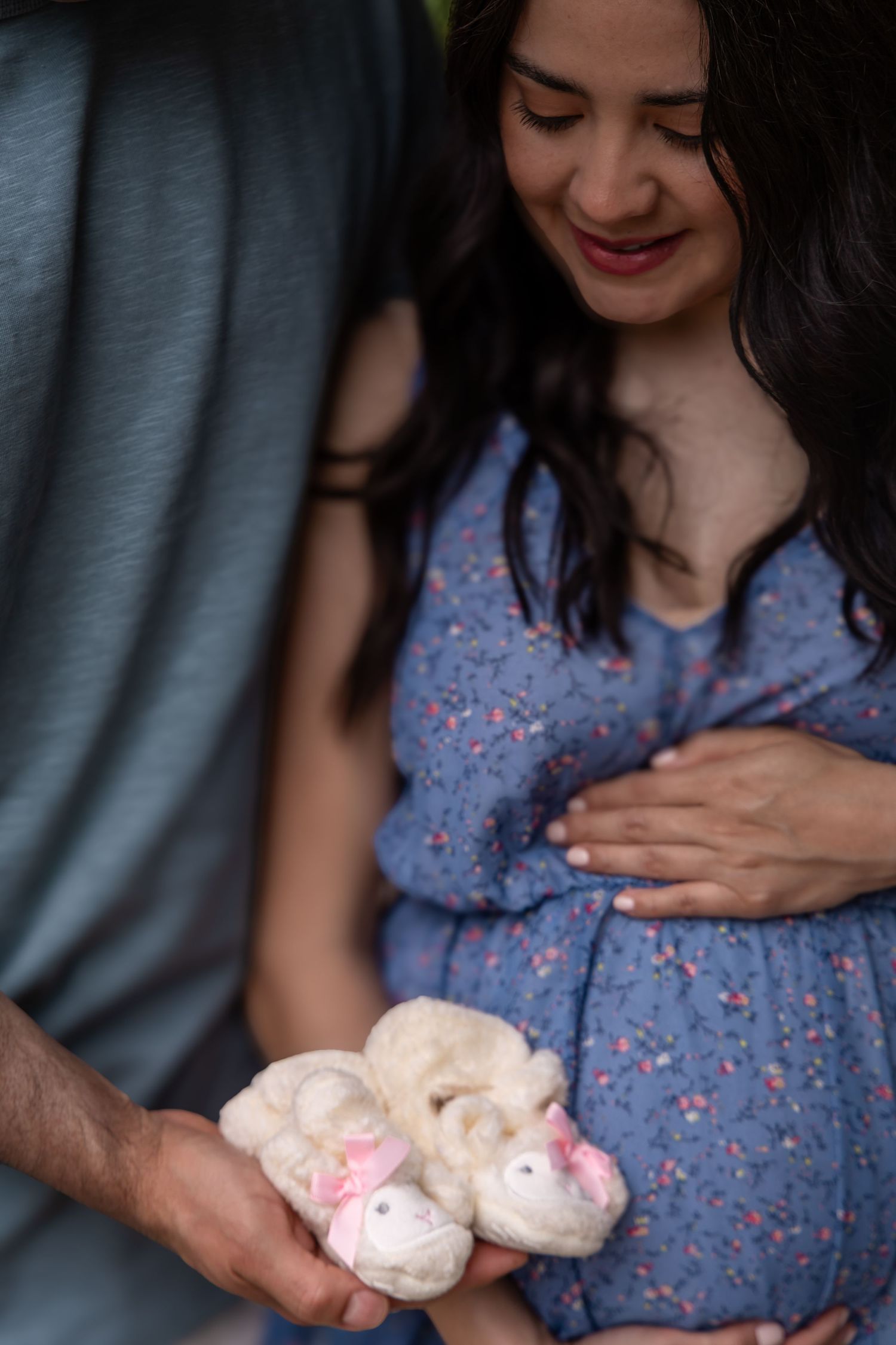 Pregnant woman in blue floral dress smiling as partner holds tiny pink baby booties during maternity photo shoot.