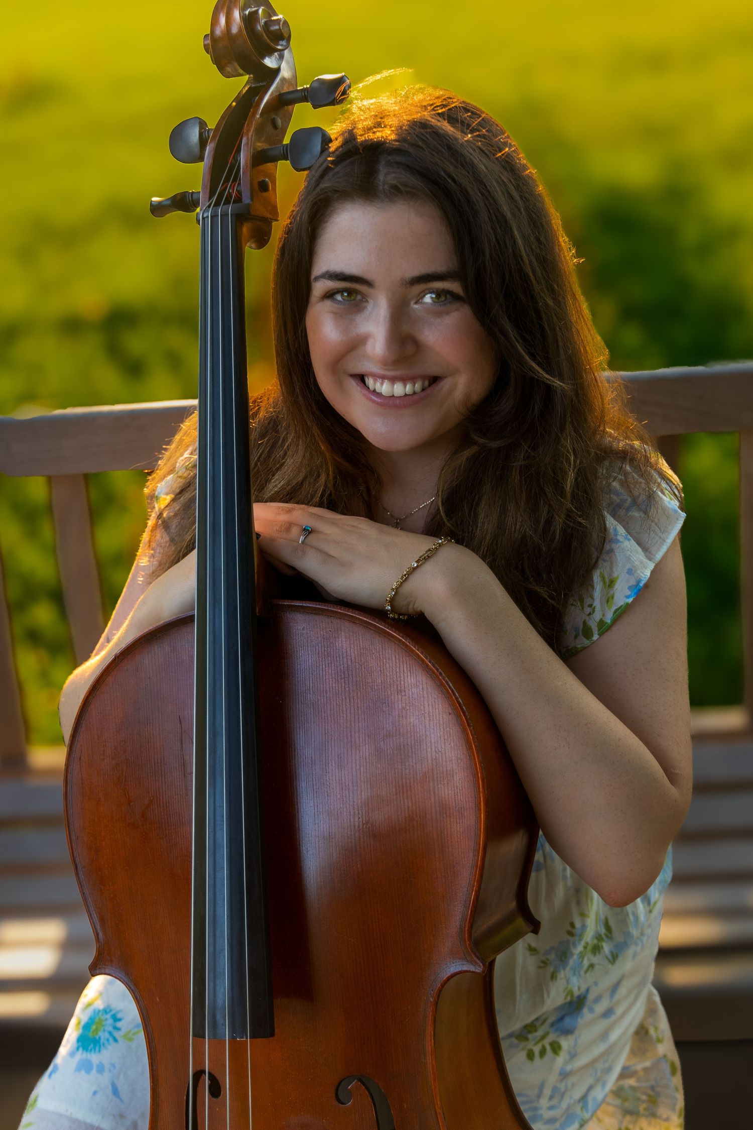 Smiling young woman with brown hair hugging a cello outdoors, with a green field in the background.